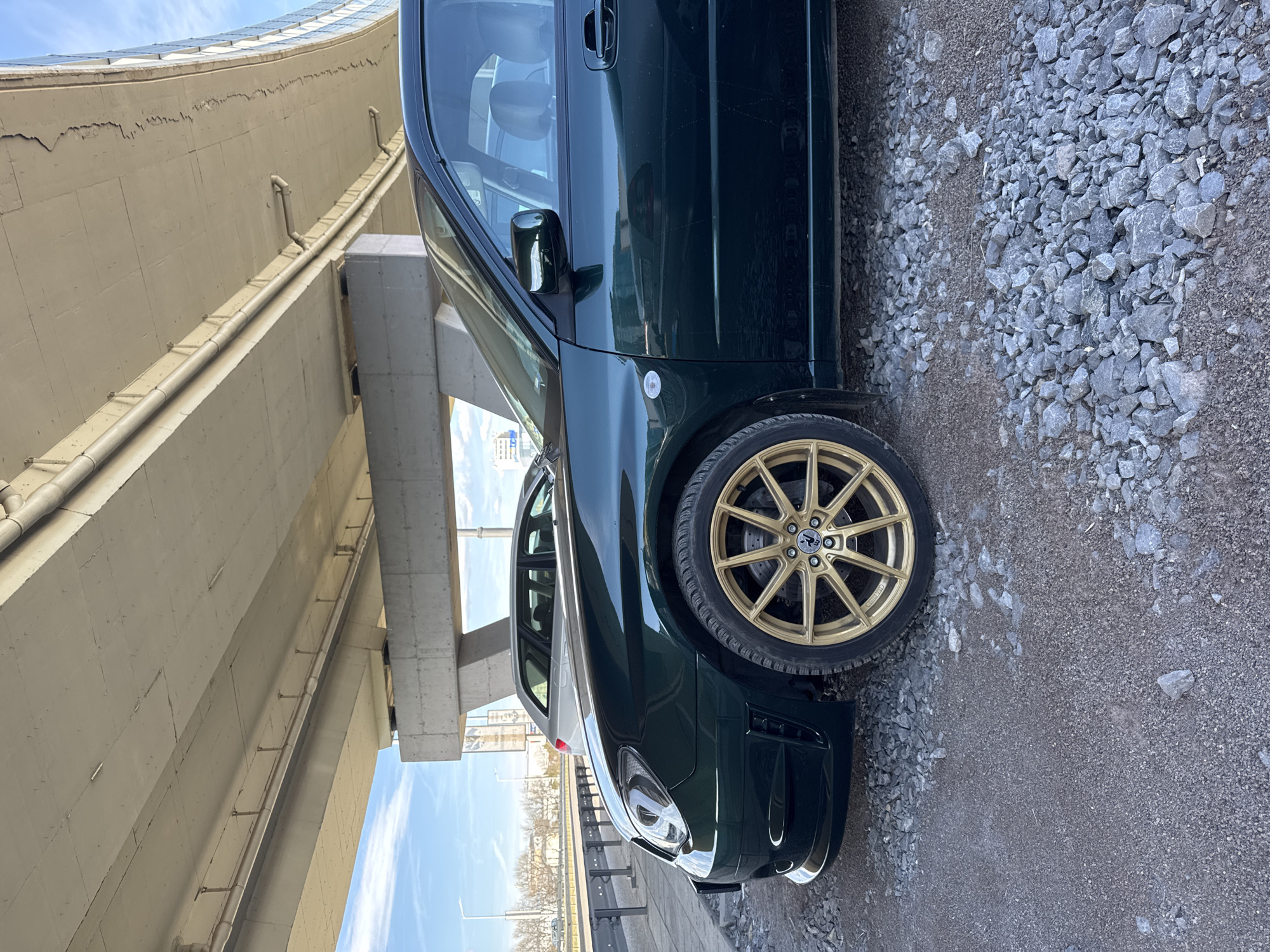 The image shows a dark green car with gold rims parked on a gravel surface underneath a large concrete bridge structure. The bridge appears to be an overpass or elevated roadway. The car is parked close to the edge of the road.
Based on the visual cues of the bridge design, guardrails, and ambient sunlight, the location is likely in Sofia, Bulgaria, specifically under one of the bridges found on the city's ring road or major boulevards.