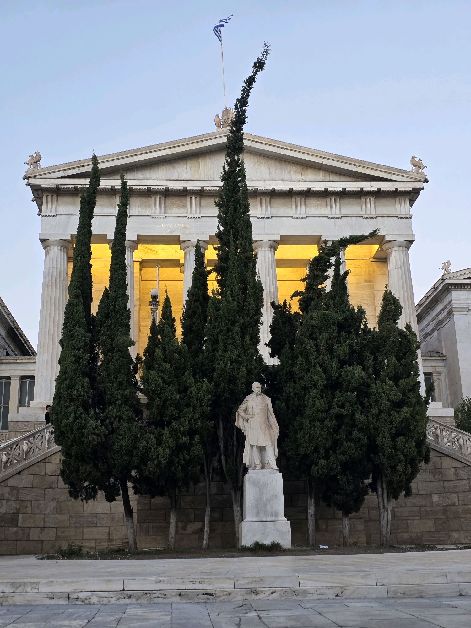 The image shows the National Library of Greece in Athens, Greece, a neoclassical building with a colonnade and pediment. In front of the building, a statue of a man in a suit stands on a pedestal, surrounded by tall cypress trees. A Greek flag flies from a flagpole atop the building. The sky is a clear, light blue, indicating it is either early morning or late evening. The lighting suggests a warm glow emanating from the interior of the building, illuminating the space behind the columns. The building's facade is made of white marble, and there are decorative elements on the roofline, including what appear to be griffins. The foreground features a paved area and stone steps leading up to the building. A person in dark clothing is visible on the steps on the left side of the image.