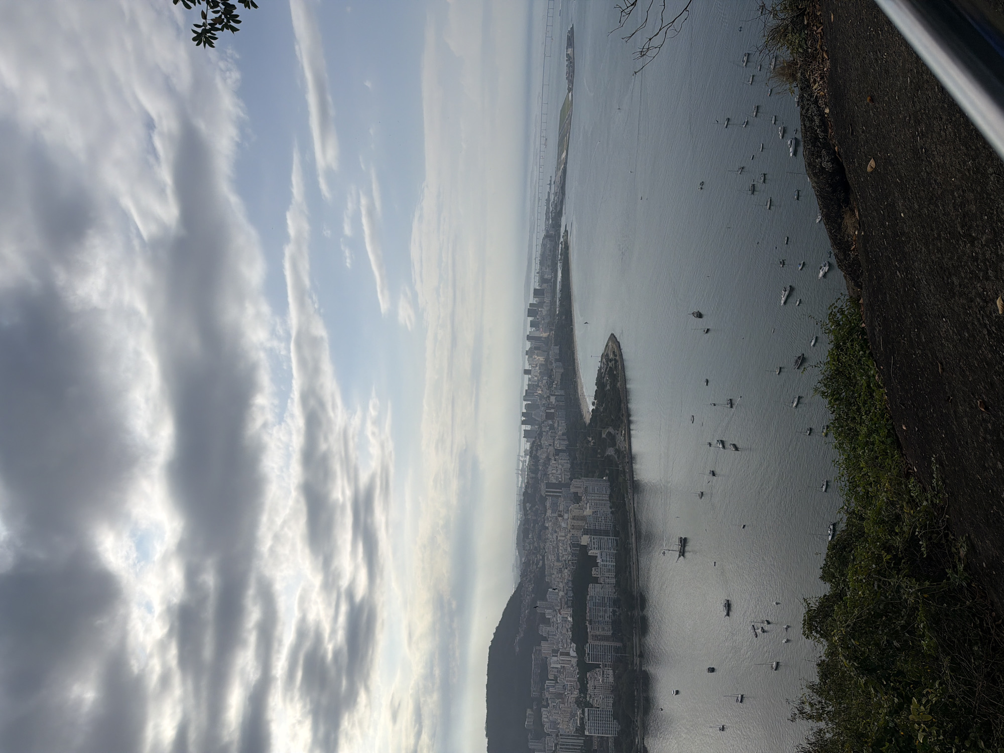 This is a high-angle, panoramic photograph of Guanabara Bay in Rio de Janeiro, Brazil, taken from a vantage point on Sugarloaf Mountain. The scene depicts a wide expanse of calm, grey water dotted with numerous sailboats and smaller vessels. Along the curve of the bay, a densely populated city skyline stretches out, characterized by a mix of modern high-rise buildings and older, lower structures. A large, dark, tree-covered mountain rises behind the city in the left portion of the frame. In the distance, across the bay, a long bridge connects to an airport runway and a series of industrial structures. The sky is overcast with dramatic grey clouds, interspersed with patches of lighter sky and a hint of sunlight breaking through. The foreground of the image is dominated by the dark, rocky terrain and lush green foliage of Sugarloaf Mountain, with a silver railing visible on the right. The overall impression is one of a vast, bustling urban landscape meeting the natural beauty of the bay and mountains under a moody sky.
