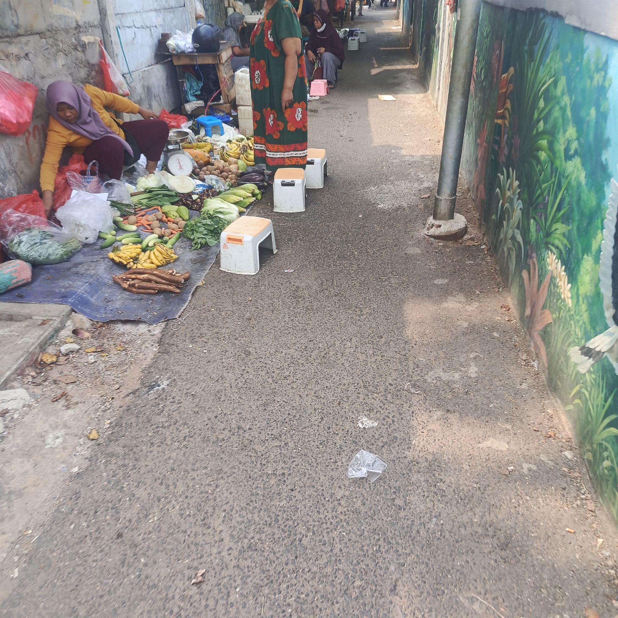 The image shows a street market scene in South Jakarta, Indonesia. A woman wearing a purple hijab and yellow shirt is seen tending to a stall selling fresh produce. The stall is set up on a blue tarp spread on the asphalt ground. Various vegetables, including leafy greens, carrots, cucumbers, corn, and bananas, are displayed. Some bananas are yellow and ripe, while others appear to be plantains or cooking bananas. A pile of cassava roots is also visible. In the background, other market-goers can be seen, and some are sitting on small plastic stools. To the right of the scene, a colorful mural depicting lush green foliage and flowers adorns a wall. A metal pole, likely part of a building structure, stands near the mural. The ground is paved with asphalt and shows signs of wear and tear, with some debris and fallen leaves scattered around. Sunlight casts shadows on the ground, indicating it is daytime.