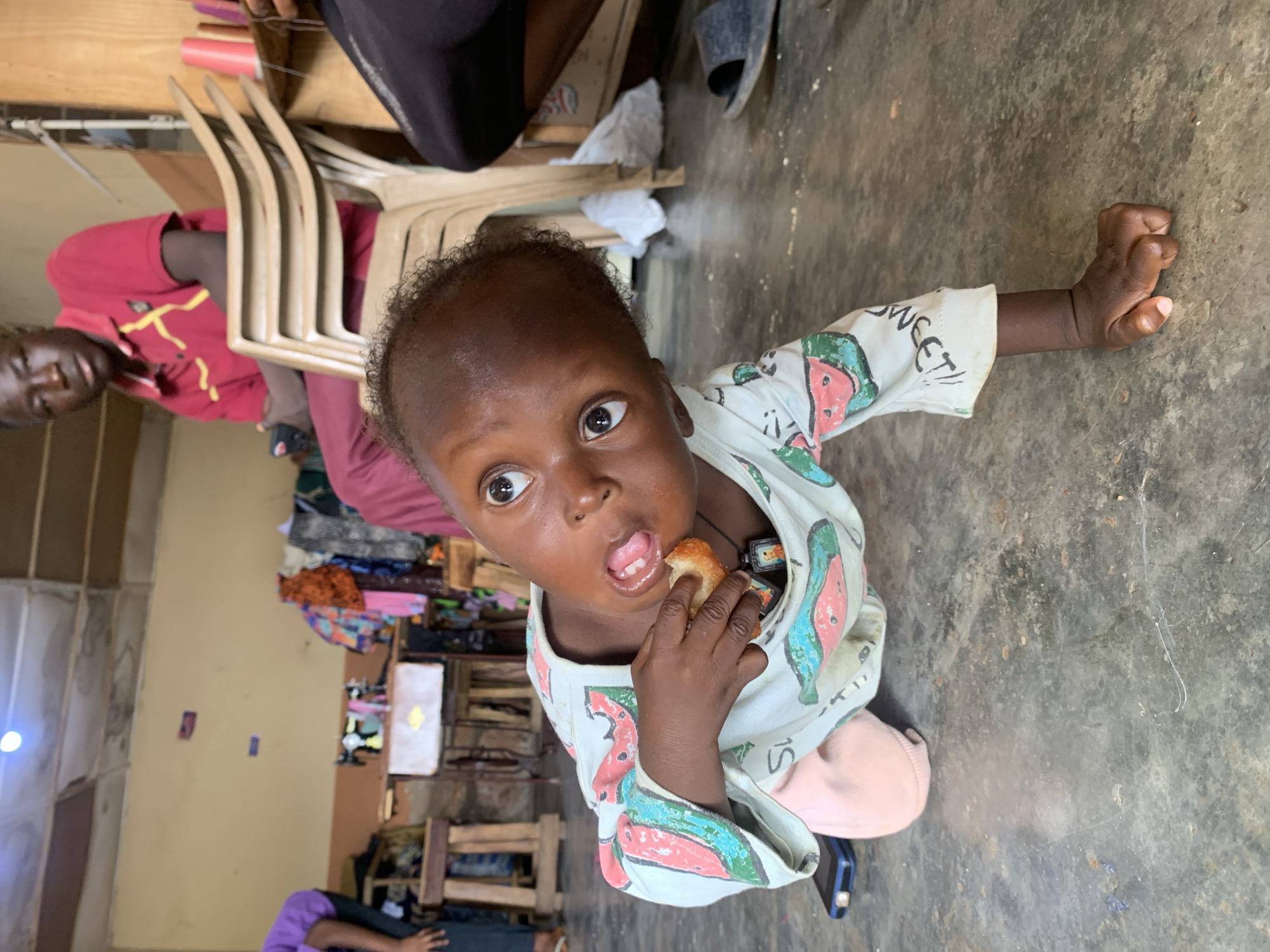 A child in Jos, Nigeria, is eating. The child has dark skin and curly hair, wears a patterned shirt, and is standing on a concrete floor. The child holds food in one hand, and extends the other arm out to the side. In the background are chairs, other people, and a wall with items attached to it.