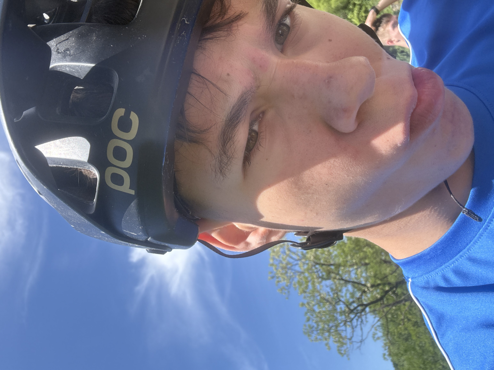A close-up, tilted shot shows a person wearing a dark-colored POC bicycle helmet and a blue athletic shirt with white trim. The person's face fills much of the frame, revealing light skin, brown hair, brown eyes, and a slight blemish on their forehead. The background consists of a blue sky with wispy clouds and a tree with green leaves partially visible in the lower right corner. Another person is partially visible in the upper right corner of the frame. Based on the visible elements, there is no information to determine whether this photograph was taken in Donchovci, Bulgaria.