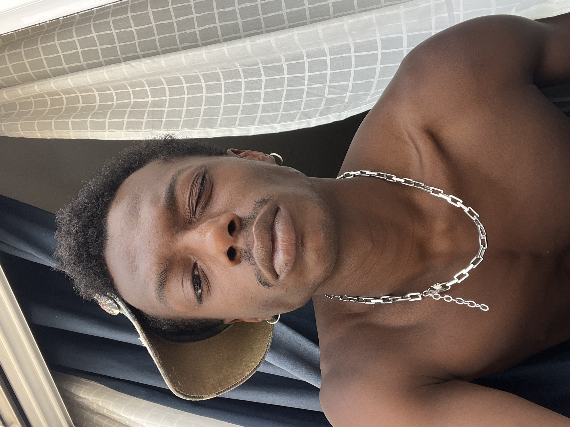 The image is a close-up selfie of a young Black man wearing a baseball cap and a silver chain necklace. He is looking directly at the camera with one eye closed in a wink. The man's skin tone is dark brown. He has short, curly dark hair and a thin mustache. He is shirtless, and his left shoulder and upper chest are visible. The background consists of dark blue curtains on the left and a white sheer curtain with a grid pattern on the right. There is a window frame visible at the top of the image. The lighting suggests it is daytime, possibly indoors near a window. There is no indication of the specific location within the city of Barcelona, Spain.