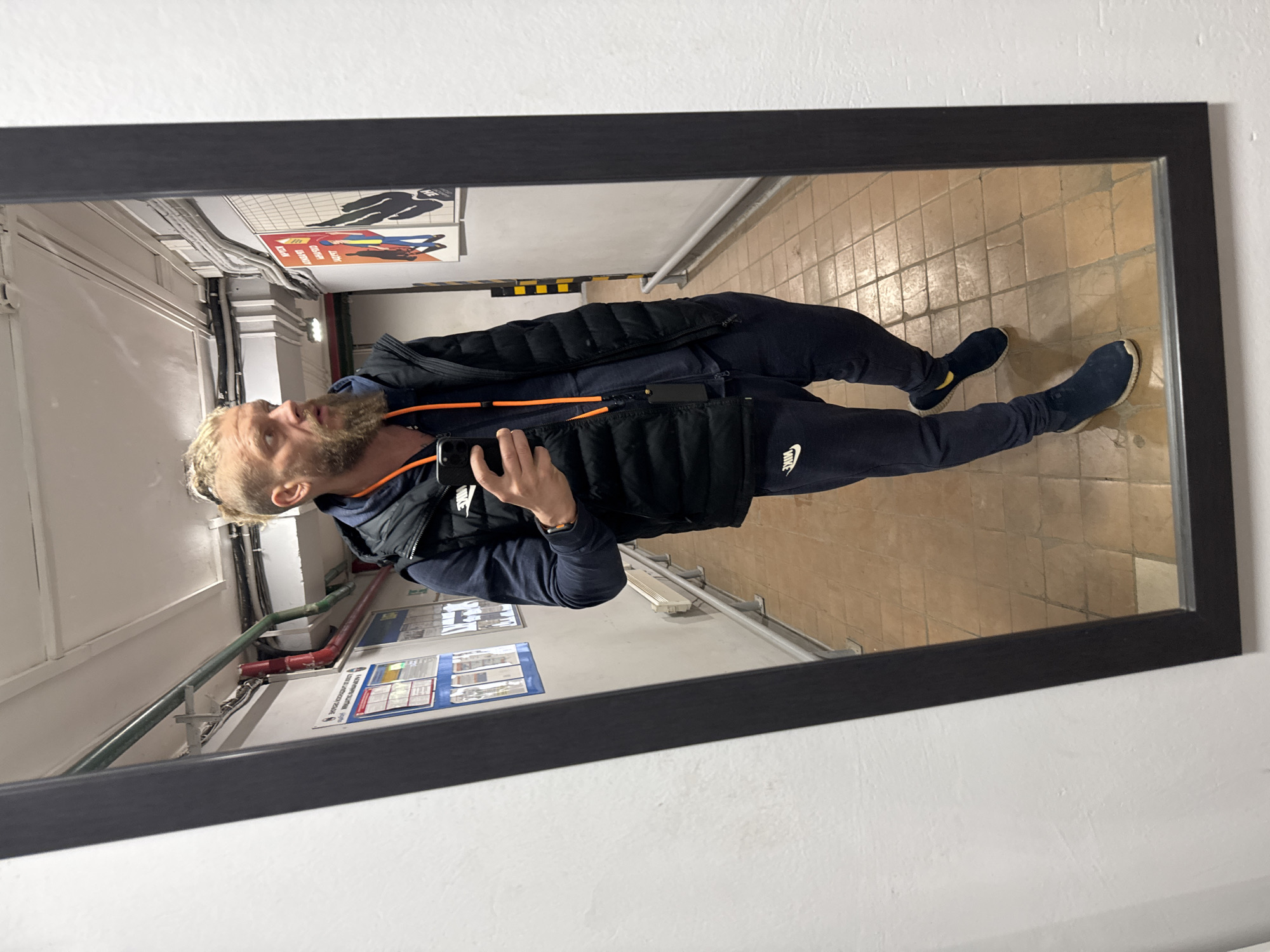 A man is reflected in a black-framed mirror. He has short blonde hair and a beard. He is wearing a dark blue long-sleeved shirt, a black puffer vest, and dark blue sweatpants with a Nike logo on the left leg. He has dark blue slip-on shoes. He is holding a smartphone in his hand. The reflection shows him standing in what appears to be a service corridor or basement area, with tiled floors and white walls. There are pipes, warning signs, and posters on the wall behind him. Based on the available details, I cannot confirm that the location is Volzhsky, Russia.