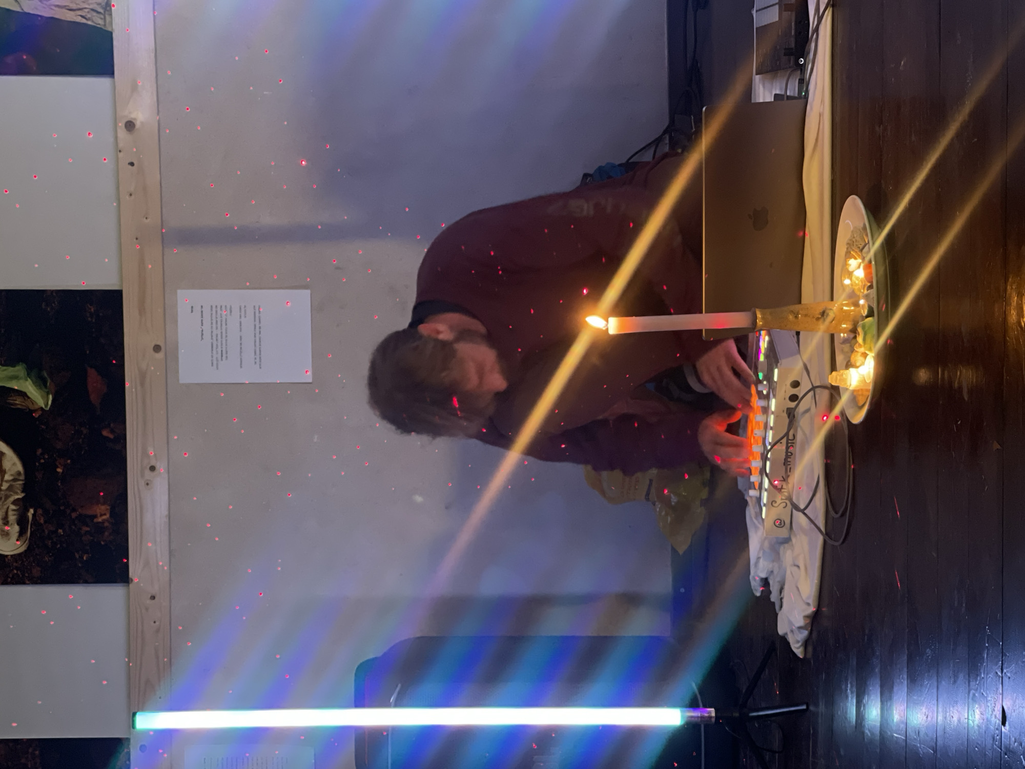 The image, originally oriented horizontally, captures an indoor artistic or performance setting. A male individual with short brown hair and a beard, wearing a dark red long-sleeved shirt, is actively engaging with electronic music equipment. His hands are placed on a white music controller or keyboard, which features illuminated buttons and sliders, indicating a performance or creative process. A silver laptop (appearing to be a MacBook) is open behind the controller, placed on a stand or surface, with a white cloth draped underneath.
The scene is dramatically lit by multiple artificial light sources. A tall, vertical LED tube light, displaying a blue, green, and purple gradient, stands on a tripod in the foreground. Numerous small red laser dots are projected across the light-colored walls and dark wooden floor, creating a starry or atmospheric effect. Prominent golden light beams, likely lens flares from a strong light source, diagonally cut through the image. A white candle in a holder is positioned near the person's hands, providing a warm, central glow. To its right, a bowl or plate holds small, warm-toned string lights.
The background features light-colored walls and a dark wooden floor. A wooden beam or frame is visible on the left wall, adjacent to what appears to be another artistic display. A sheet of paper with text, partially obscured and largely unreadable, is taped to the wall; the word "NOTE" is faintly visible. The word "MUSIC" is also discernable on the white electronic controller. The elaborate lighting and equipment setup suggest an art installation, a live electronic music performance, or a similar creative event, likely held indoors in the evening or at night. The location context is Mitzpe Ramon, Israel.