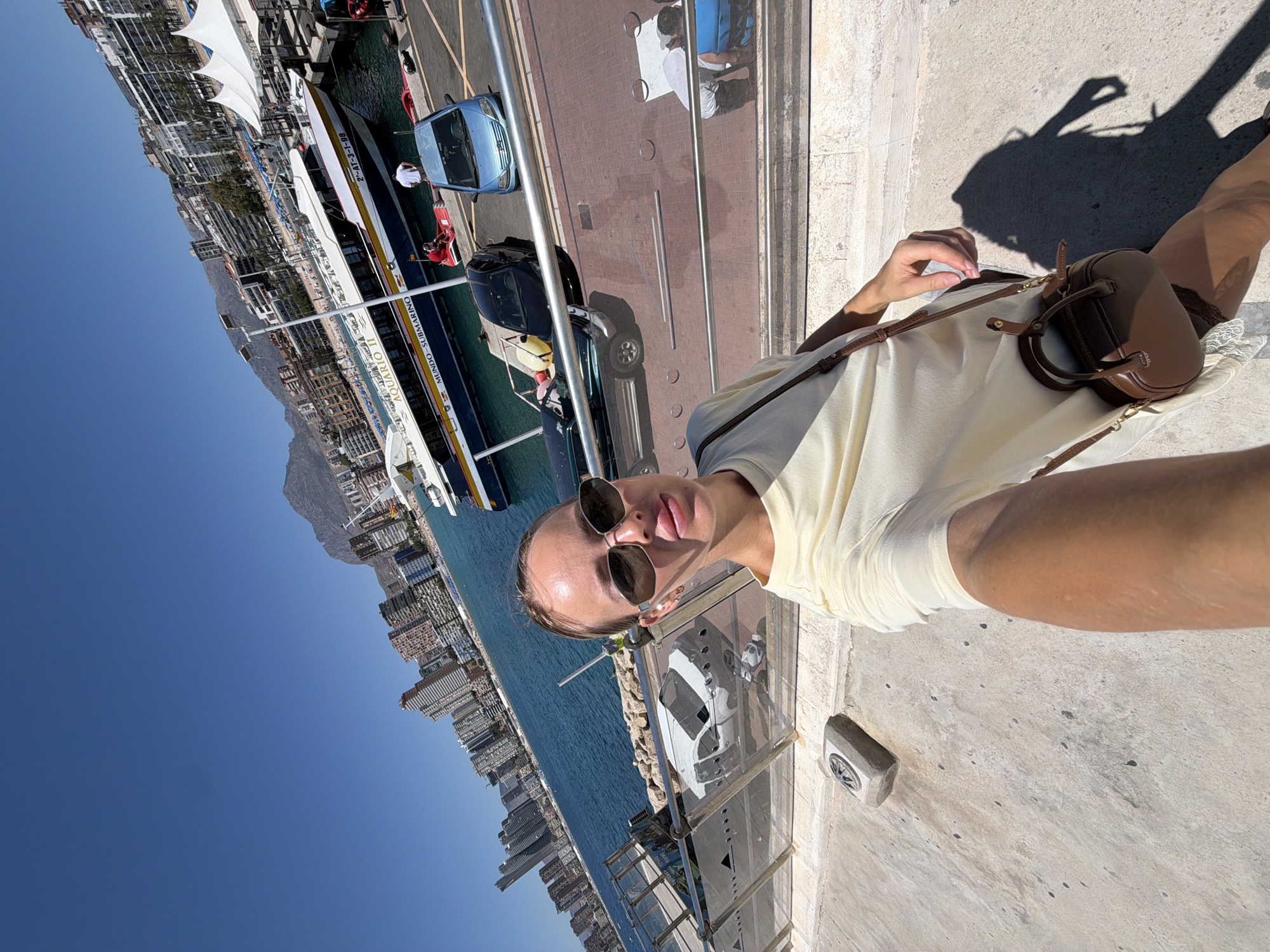 A woman is taking a selfie outdoors, standing on a concrete walkway with a railing. She is wearing a light yellow dress and sunglasses. A small brown purse is slung across her body. Behind her, a body of water, a beach with buildings, and mountains are visible under a clear blue sky. Several boats are docked in the water, and cars are parked along the walkway. The scene appears to be the port area of Benidorm, Spain, known for its high-rise buildings and beaches.