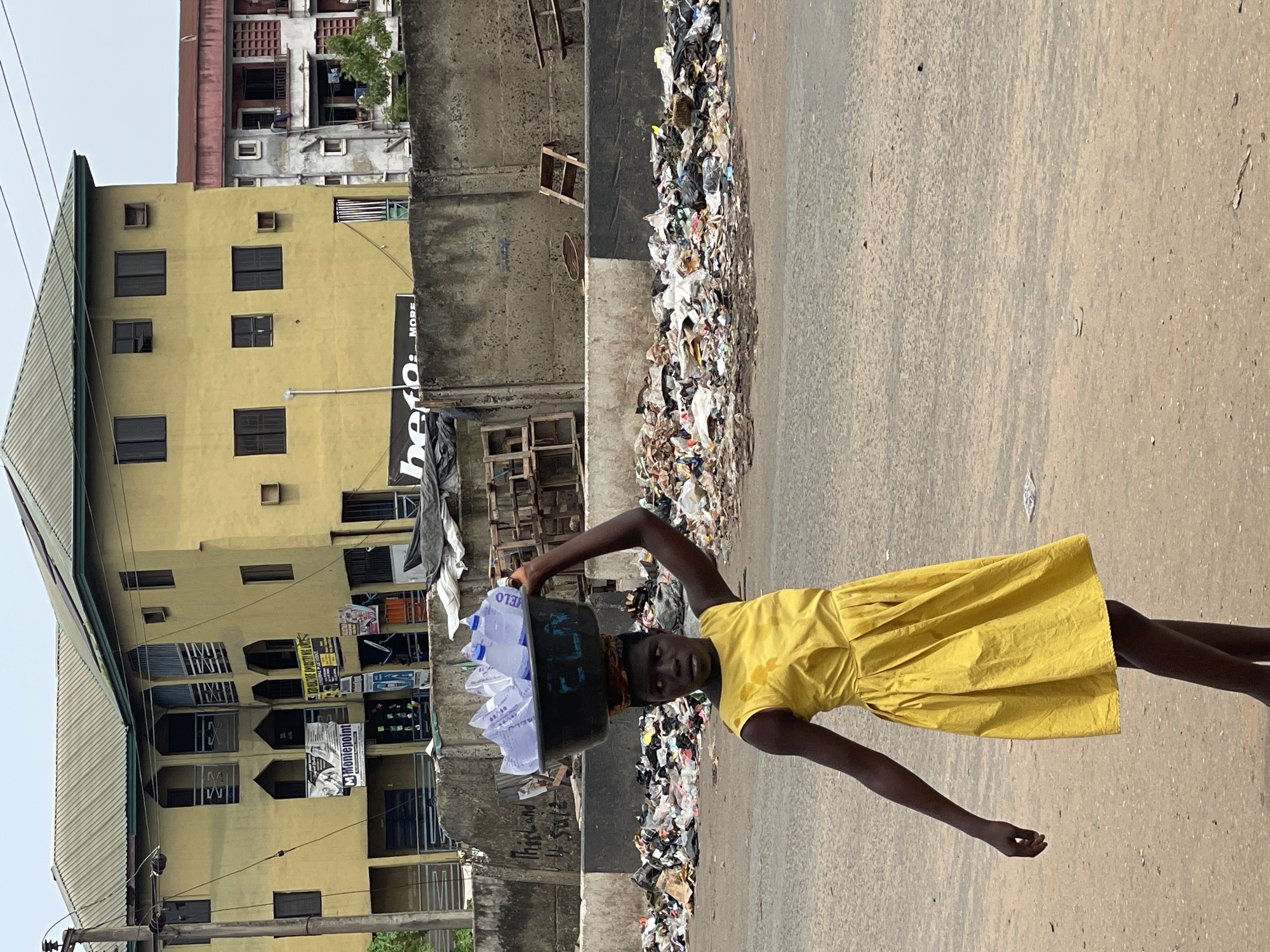 A young woman in a yellow dress is carrying a black bucket filled with water sachets on her head. She stands next to a large pile of rubbish on a street in Onitsha, Nigeria. The background shows a yellow building with several windows and posters, adjacent to a concrete wall.