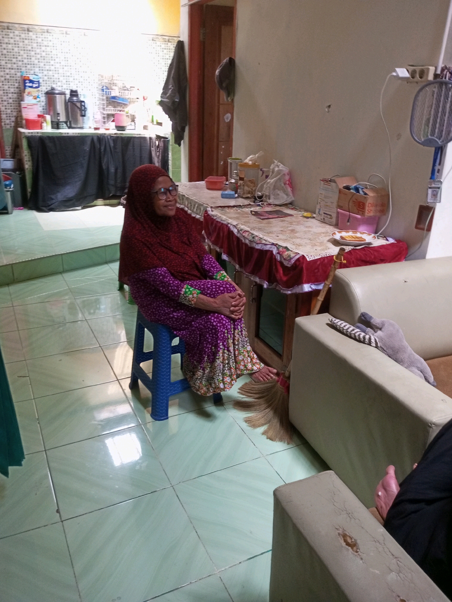 An elderly woman wearing glasses and a maroon hijab sits on a blue plastic stool. She is dressed in a long, purple, polka-dotted dress with green accents at the sleeves and hem. The woman is seated in what appears to be a domestic setting, likely a kitchen or dining area in Krasak, Indonesia. The floor is tiled in a light green color. To her right, a wooden table is covered with a patterned tablecloth. On the table are various items, including plates, jars, and boxes. A broom is visible near the table. In the background, a counter with kitchen appliances like thermoses and a pink kettle can be seen. A black cloth drapes over part of the counter area. To the far right of the image, a light-colored sofa is visible. A hand is resting on the sofa's armrest, and a gray stuffed toy is draped over another part of the sofa. The overall impression is a candid snapshot of a woman in a home environment.