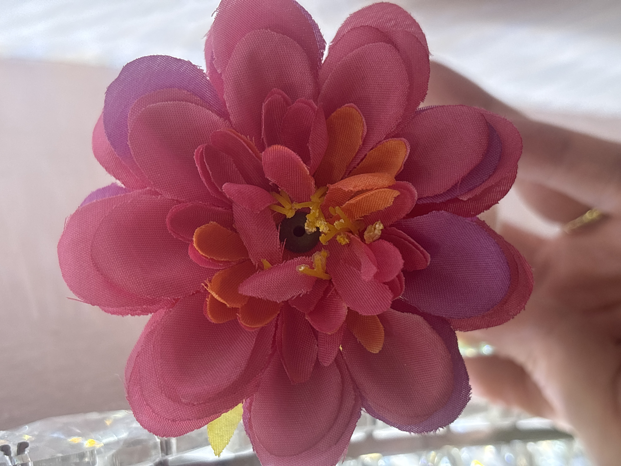 The image presents a close-up view of a vibrant artificial flower, likely crafted from fabric, positioned centrally in the frame. The flower features numerous layered petals, primarily in shades of pink and fuchsia. Towards the center, smaller petals exhibit warm orange and yellow tones, while some of the outermost petals introduce hints of purple. The very core of the flower contains a dark, bead-like element surrounded by delicate yellow stamen-like structures. The fabric texture of the petals is visible, with some edges appearing slightly frayed.
The background is softly blurred, creating a shallow depth of field. On the right side of the image, part of a human hand with fair skin is subtly visible, appearing to hold or support the flower. A faint metallic glint, possibly from jewelry, can be observed on the hand. Below the flower, the out-of-focus area suggests a reflective surface, potentially glass or clear plastic, where scattered light reflections and bokeh effects, including some yellow-golden highlights, are discernible.
The lighting is bright and even, suggesting a daytime setting. There is no specific activity or event taking place other than the presentation of the flower. No text is visible, and there are no discernible location cues to indicate Sofia, Bulgaria, or any specific outdoor or indoor environment beyond a general bright setting. The image focuses on the intricate details and colors of the artificial bloom.