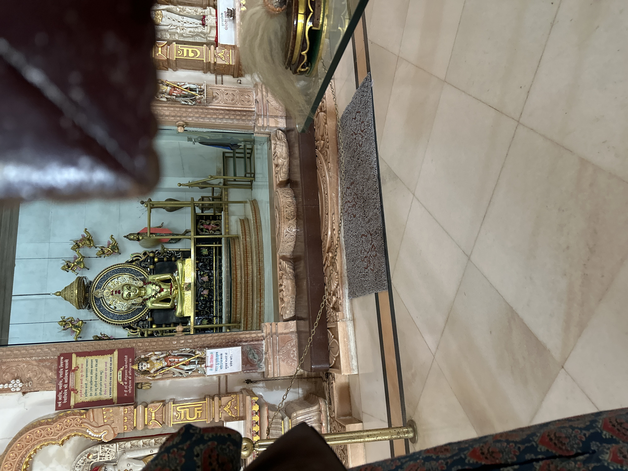 The image shows a view of the interior of a temple. In the background, a golden statue of a deity is seated on a raised platform, adorned with intricate carvings and a decorative umbrella. The statue is positioned centrally within the temple's main altar area. Flanking the altar are ornate pillars and smaller statues. In the foreground, a patterned rug is placed on a tiled floor. A glass table with a decorative item on it is visible on the right side of the frame. The overall environment suggests a place of worship. The provided location context indicates this scene is from Shahapur, India.
