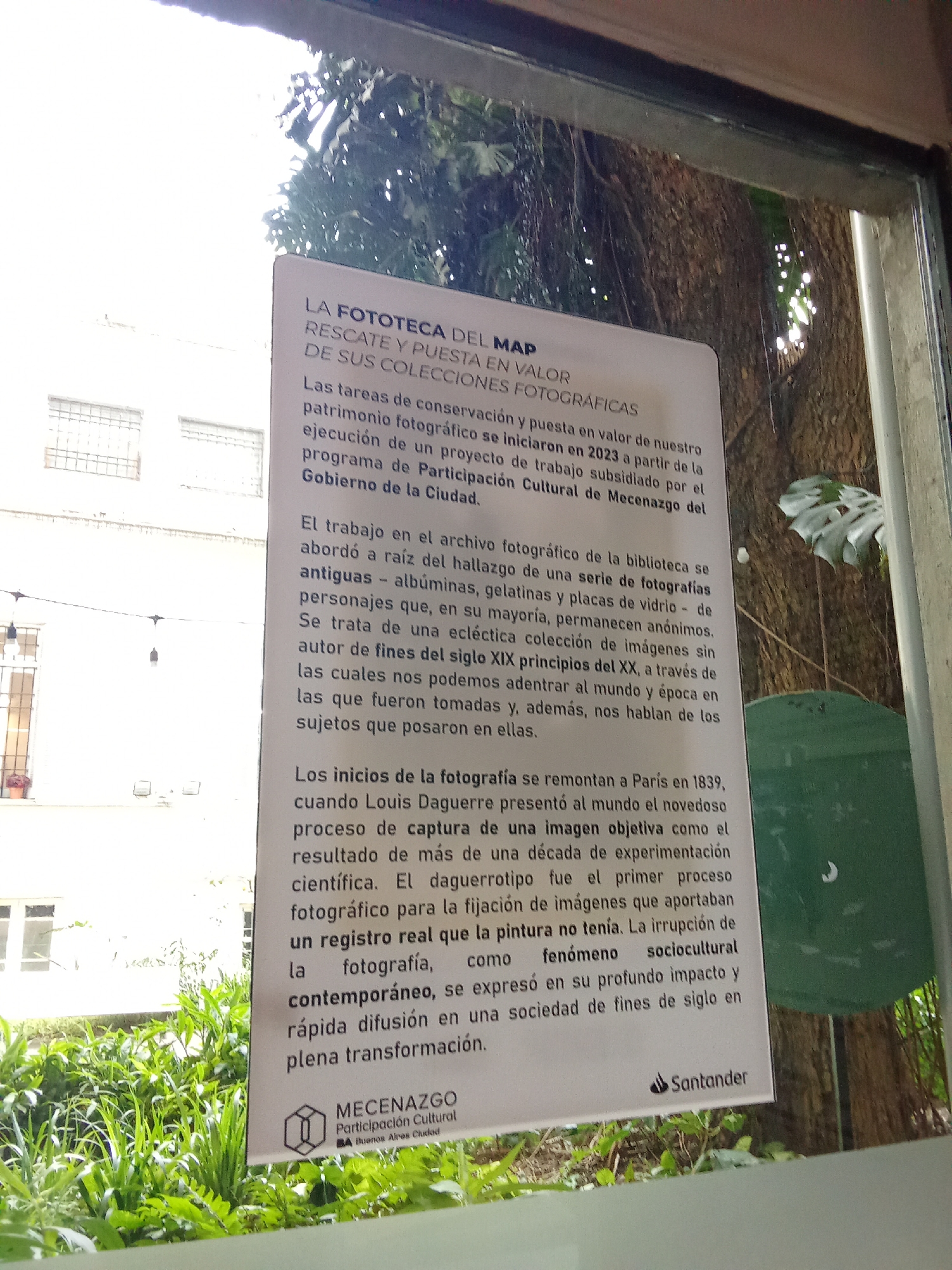 The image shows a sign displayed on a window or glass surface, with a green, leafy background visible through the glass. The sign itself is white with black text. The top of the sign reads "LA FOTOTECA DEL MAP RESCATE Y PUESTA EN VALOR DE SUS COLECCIONES FOTOGRÁFICAS". The sign then elaborates on the photographic collection, stating that its preservation and enhancement began in 2023 as part of a work project subsidized by the "Programa de Participación Cultural de Mecenazgo del Gobierno de la Ciudad". It details that the work in the photographic archive of the library began after finding a series of photographs, including albums, gelatin plates, and glass plates, most of which are anonymous. It describes a collection of images from the late 19th and early 20th centuries that allow us to understand the world and the era through the subjects captured. The sign also touches on the origins of photography, mentioning Paris in 1839 and Louis Daguerre's presentation of an objective image as a result of experimentation. It highlights the daguerreotype as the first process for fixing images that painting could not achieve, and describes the advent of photography as a sociocultural phenomenon with a profound impact on late 19th-century society. At the bottom of the sign, a logo for "MECENAZGO Participación Cultural BA Buenos Aires Ciudad" is visible, along with the Santander logo. The outdoor environment behind the glass consists of dense foliage and trees, with sunlight filtering through the leaves.