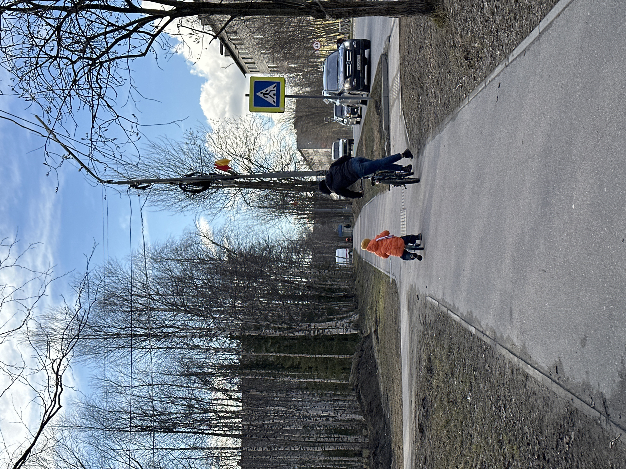 A person on a bicycle is riding down a sidewalk with a child on a scooter. There is a pedestrian crossing sign visible and several cars on the adjacent street. Trees border the sidewalk. The sky is blue with clouds. The location is unknown, but appears to be an urban or suburban area.