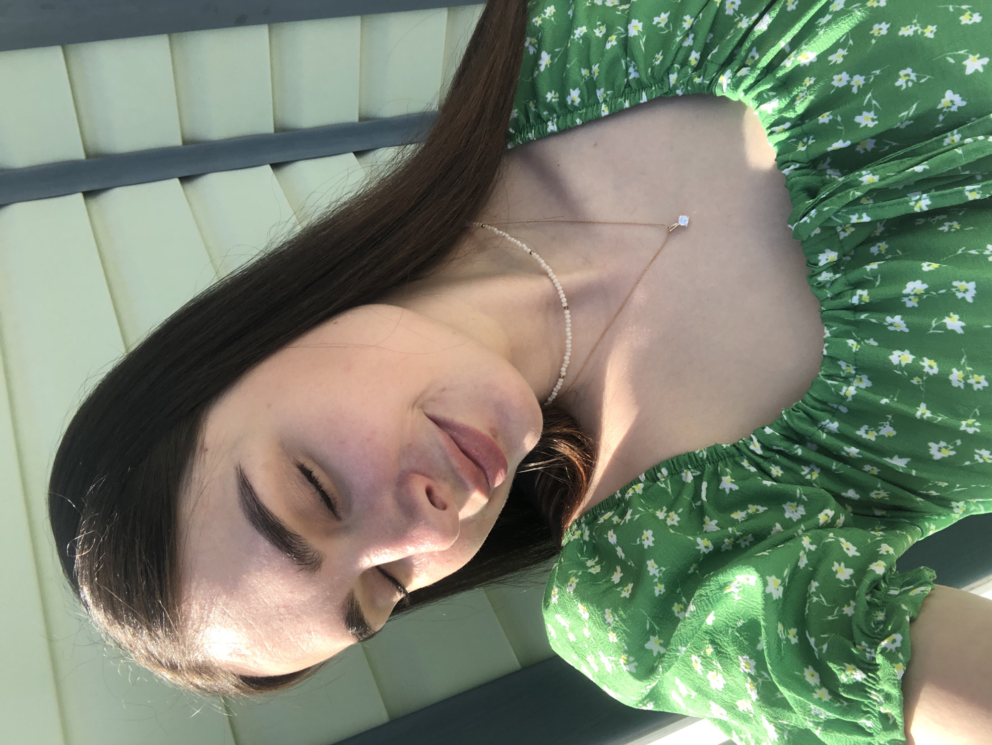 A young woman with long, straight brown hair is captured in a medium-close shot. She is wearing a green dress with a white floral pattern and a pearl necklace with a small pendant. Her eyes are closed, and she appears to be serene. The background shows upholstered panels. The location is not apparent.