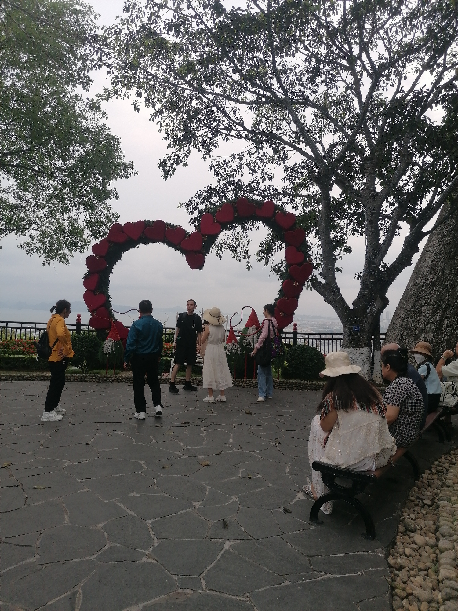 Captured in Hong Gai Ward, Vietnam, the scene unfolds outdoors on a cloudy day. A group of individuals, including sightseers, gather near a sizable heart-shaped structure adorned with red hearts. To its sides are garden gnomes. One group of individuals is standing near this structure, while another group of people is sitting on a nearby bench. The area is paved with flagstones. The location provides an elevated view with a fenced balcony visible in the background. Foliage, trees, and parts of the city are visible.