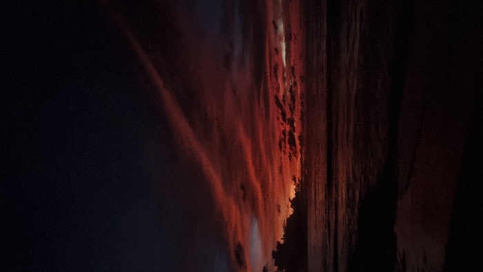 The scene depicts a dusk ocean view in Pecatu, Indonesia. The sky exhibits a dominant gradient of intense red and orange hues near the horizon, transitioning to dark blue and black in the upper regions. Multiple elongated, red-tinged cloud streaks extend diagonally across the top-right portion of the sky, with other darker, irregular cloud formations present along the horizon. The ocean surface is dark, reflecting the twilight conditions, with discernible wave patterns extending towards the foreground. The immediate foreground consists of a dark, indistinct stretch of shoreline. No individuals, specific objects, actions, or interactions are visible.