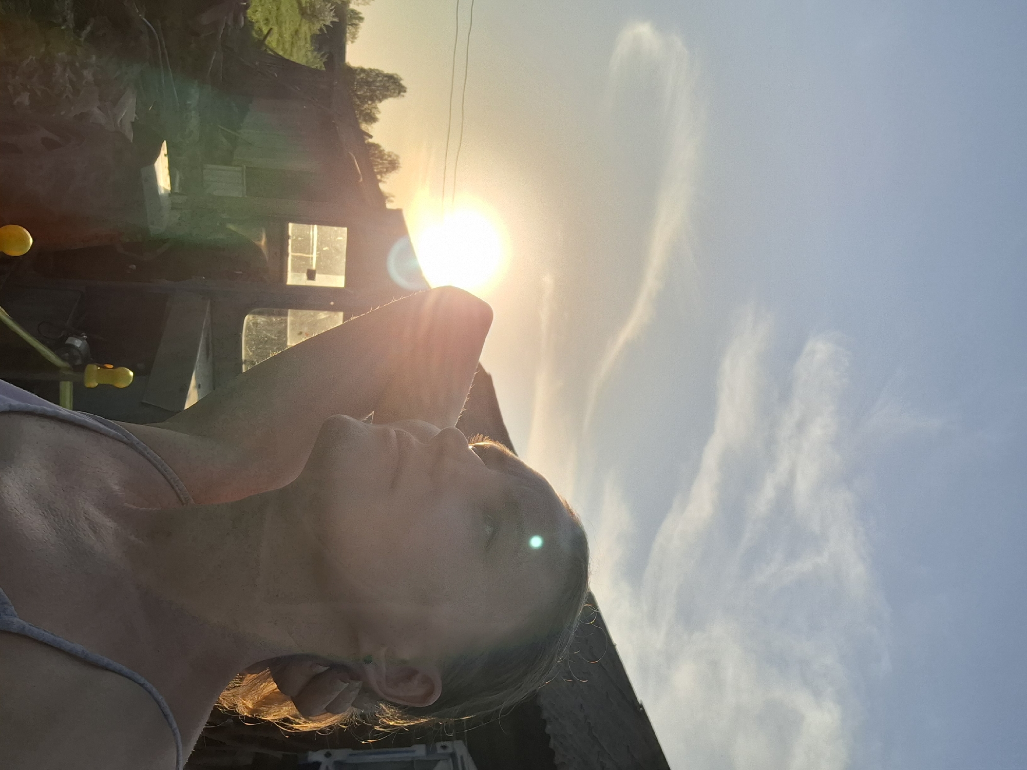 A low-angle, rotated outdoor photograph depicts a person against a bright sun and sky. The person, likely female, is fair-skinned with their head tilted back. Her hair appears to be blonde, catching the sunlight. Her left hand (as viewed) is raised behind her head. She is wearing a light-colored sleeveless top.
In the background, blurred shapes suggest buildings or structures, possibly rural or agricultural. There's vegetation present and a hint of yellow objects hanging off the structures. The sky is a gradient from light yellow to blue.
It is not possible to confirm the location of the city of Шешурга, Russia as there are no explicit landmarks or signs that would indicate a precise location. The scene depicts an individual relaxing outdoors, perhaps in a rural or semi-rural setting, during golden hour.