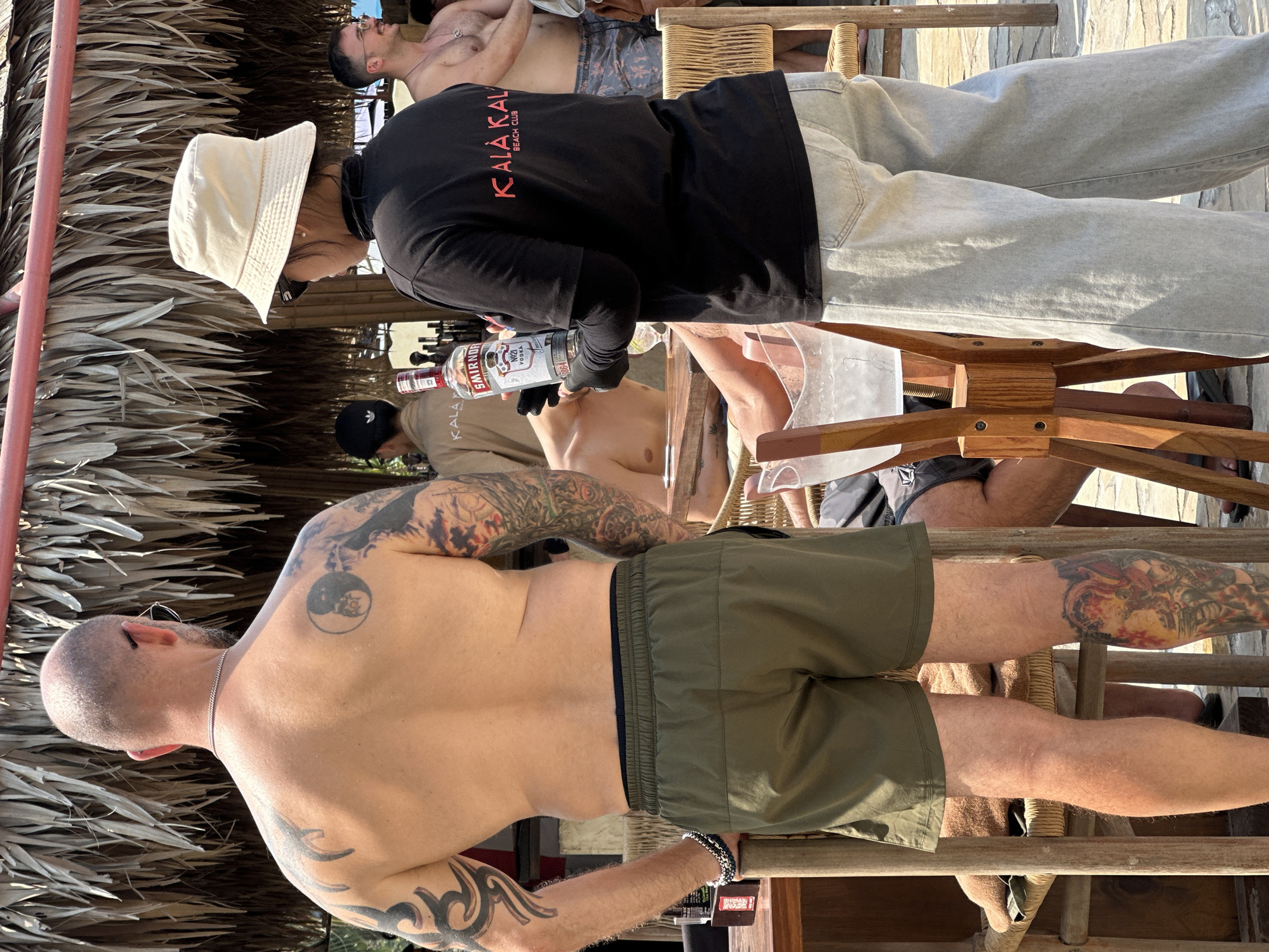 The image depicts a partially covered outdoor setting, possibly a beach bar. At least four people are visible. A man with a shaved head and extensive tattoos on his back, arms, and legs is seated in a chair. Another person, wearing a black shirt with "KALA KALA BEACH CLUB" printed in red, along with light-colored pants and a white bucket hat, appears to be serving a clear liquid from a Smirnoff vodka bottle. There are other individuals in the background, one shirtless lying down on a lounge chair. The scene appears to be at a beach club, likely in a tropical or coastal setting. Based on the shirt, the location is Đà Nẵng, Vietnam.