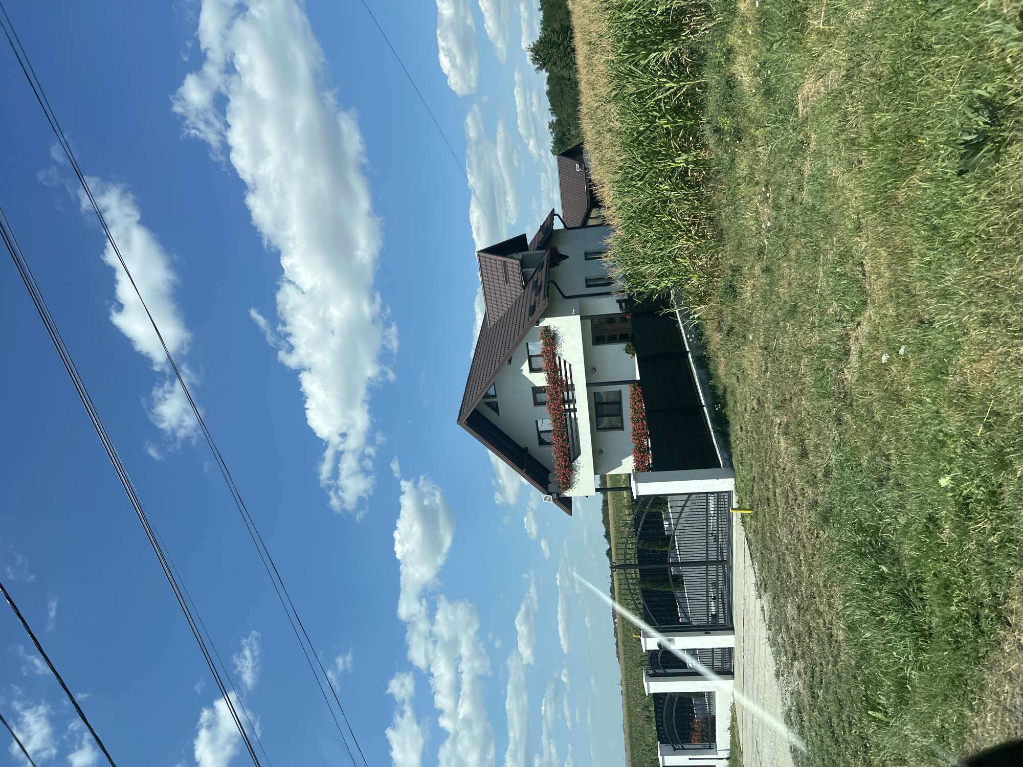 A white, two-story house with a brown, pitched roof is situated behind a black metal fence and a white-pillared gate. The house has a balcony on the second floor adorned with red flowers in planters. Adjacent to the house, a field of tall green corn stalks stretches into the background. The sky above is a clear blue with scattered white cumulus clouds. Power lines are visible crossing the upper portion of the image. The scene appears to be in the rural area of Sasca Nouă, Romania, given the presence of agricultural land and a detached residential structure. The ground in the foreground is covered with dry grass and some patches of green.