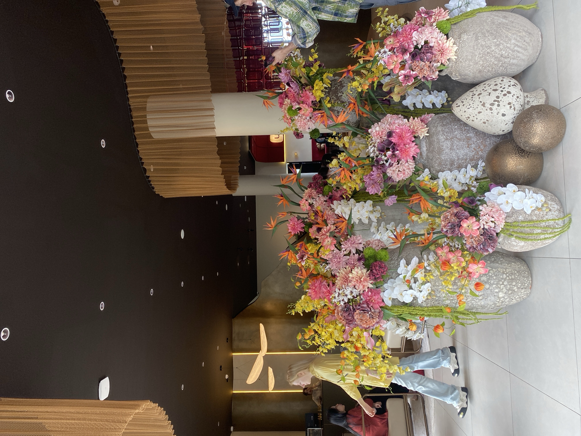 The image depicts an indoor scene, likely a lobby or public space, filled with large, decorative floral arrangements. The ceiling is dark and features numerous recessed spotlights. Hanging from the ceiling on the left and curving around a pillar on the right are gold chain-like decorative elements. In the foreground, several large, textured vases hold an abundance of colorful artificial flowers in shades of pink, yellow, orange, and white, with some greenery. Two of the vases are flanked by decorative golden orbs and a speckled white mushroom-shaped object.
In the mid-ground, a young woman with blonde hair, wearing a yellow top and light blue jeans, walks past the floral display. To her left, another woman with dark hair, wearing a red top, is seated. In the background, behind a counter, a person is visible, and further back, the interior of the building is partially seen with red seating and architectural details. On the right side of the image, a person wearing a plaid shirt and a blue cap is taking a photograph of the floral arrangement with a smartphone. The floor is tiled with large, light-colored square tiles. The overall impression is of an elegantly decorated interior space. There is no information in the image to indicate the location in Da Lat, Vietnam.