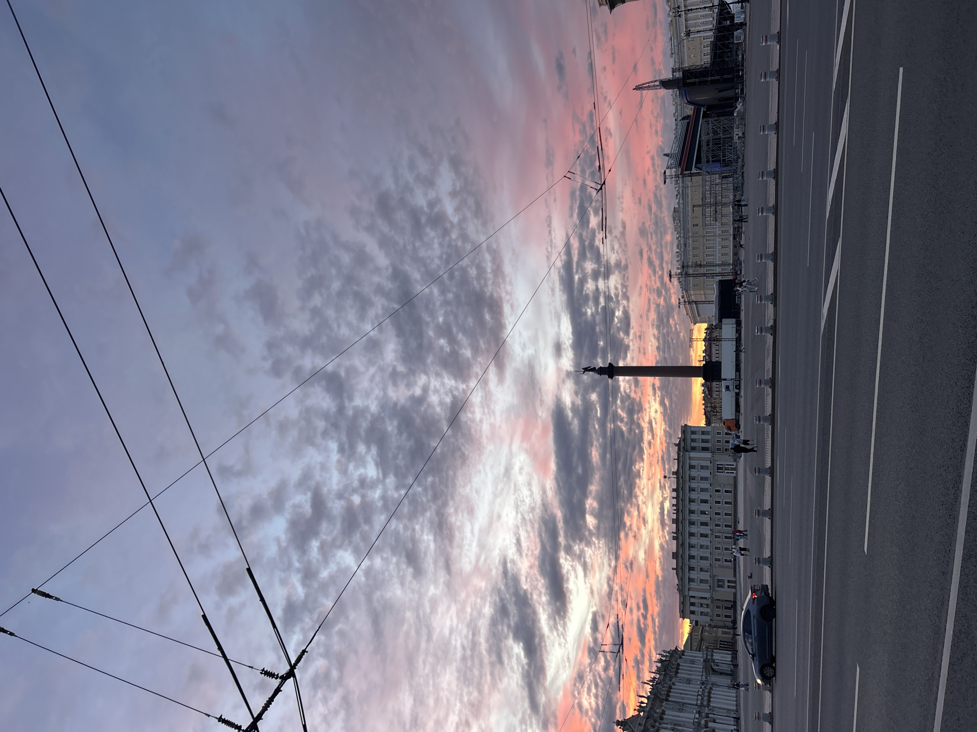 The image depicts a city square at dusk, with a dramatic sky painted in hues of pink, purple, and gray. The foreground features a multi-lane road with white markings. In the middle ground, a wide pedestrian area is bordered by a row of white bollards. Several people are scattered across the square, walking and standing. Dominating the scene are large, classical-style buildings lining the perimeter of the square. To the left, a grand, ornate building with many windows is visible. In the center, a tall column topped with a statue stands prominently. To the right, another large building complex is under construction or renovation, with scaffolding and large structures partially covered. Overhead, a network of electrical wires crisscrosses the sky, obscuring parts of the view. A dark-colored car is parked on the left side of the square, near the road. The overall atmosphere suggests a public gathering space in an urban environment, captured during the twilight hours.