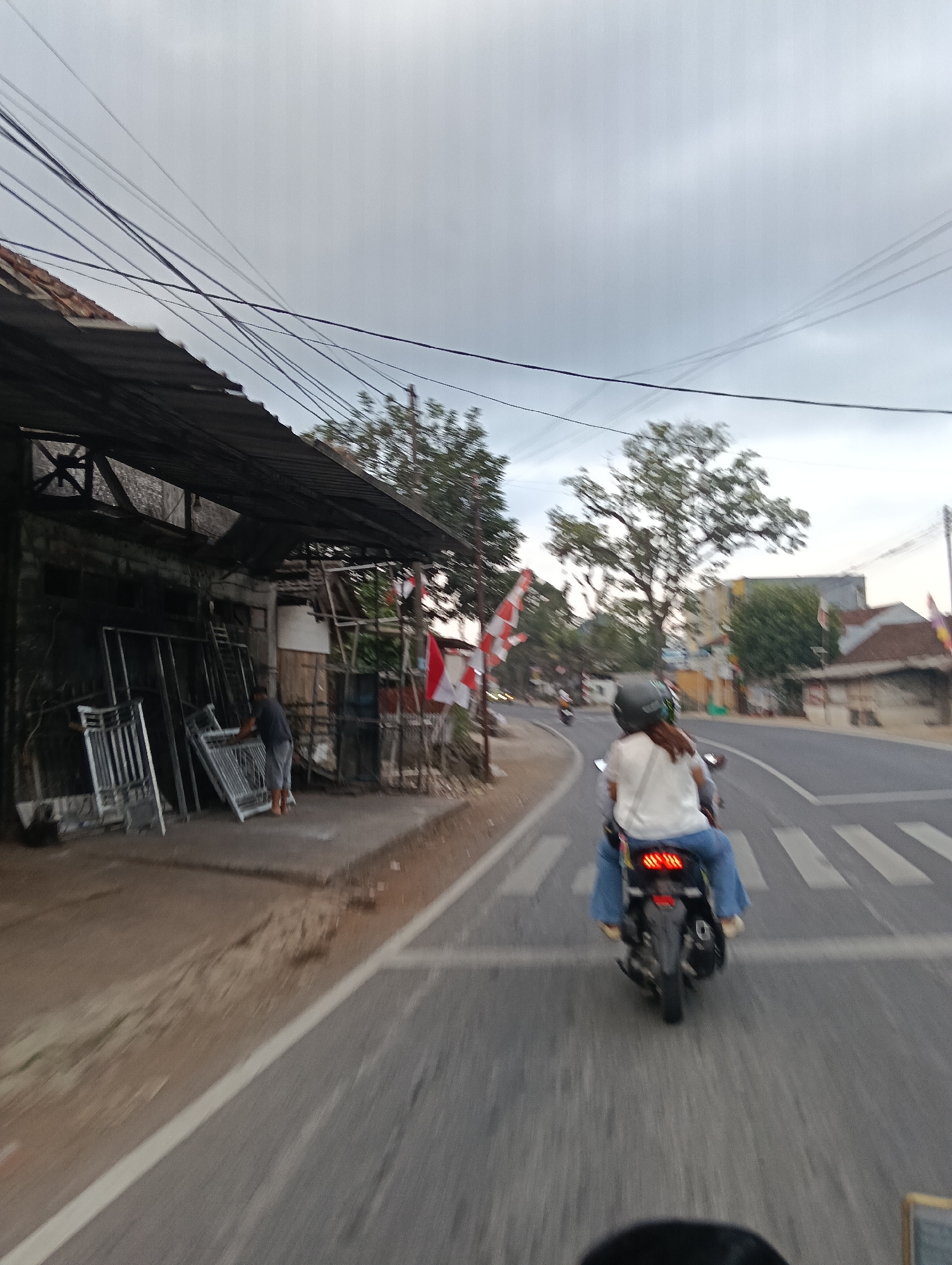 This is a slightly motion-blurred, eye-level, outdoor, daytime photograph taken from the perspective of someone on a motorcycle, looking forward. The scene depicts a street in what appears to be Cintaraja, Indonesia.
In the foreground, the rear of a motorcycle is visible, with a person wearing a white t-shirt and blue jeans riding it. They have a dark helmet on. The motorcycle is crossing a pedestrian crossing.
To the left of the road, there is a building with a dark, corrugated metal roof supported by wooden beams. In front of the building, metal frames and structures are stacked. A person wearing a dark shirt and grey shorts is standing and appears to be interacting with one of these metal structures. Indonesian flags, red and white, are visible on poles near the building.
The road itself is paved and has white lane markings. Beyond the road, there are trees and buildings. The sky is overcast, suggesting cloudy weather. Power lines are strung across the upper portion of the image.
