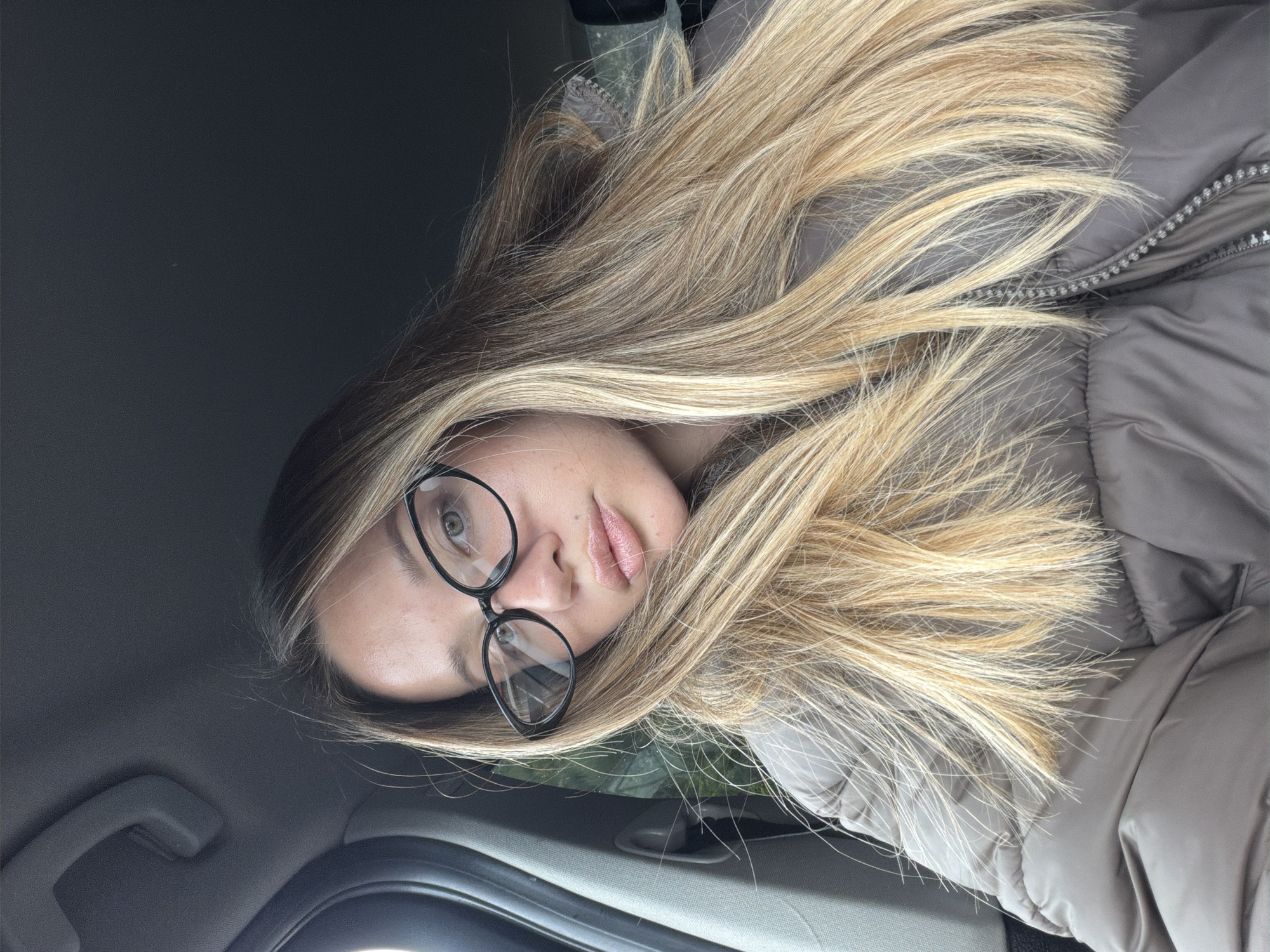 This image depicts a female individual seated in the passenger side of a vehicle within the city of Unknown, Unknown. The subject has long, multi-toned blonde hair, light-colored eyes, and is wearing black, cat-eye style eyeglasses. Her lips are slightly parted and appear to have a reflective coating. She is wearing a dark taupe or brown padded jacket, partially zipped, with the collar visible. The vehicle's interior features a dark gray headliner and a matching grab handle visible in the upper left quadrant. The external environment through the window on the left is out of focus and indistinct. The individual is looking directly at the camera.