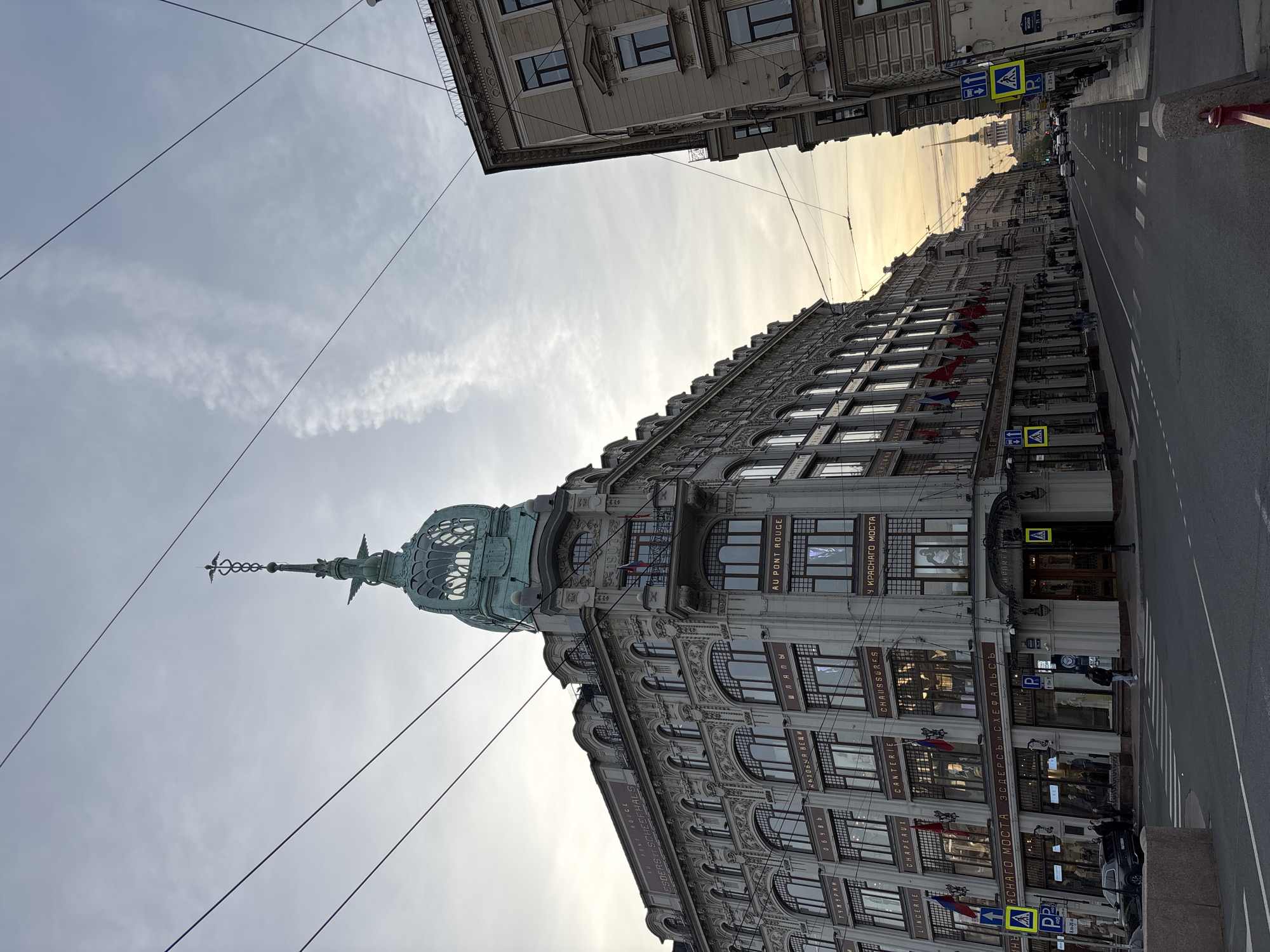 The image shows a view of the "Au Pont Rouge" building in Saint Petersburg, Russia, from a low angle, looking upwards. The building features a distinctive tower with a spire topped by a caduceus. The building's facade is adorned with multiple windows. The street stretches along the right side of the image and a building can be seen across the street to the left. Overhead wires cut across the sky. Street signs with crosswalk markings are present. The sky is light and partly cloudy. The building's name is visible in both French and Russian.