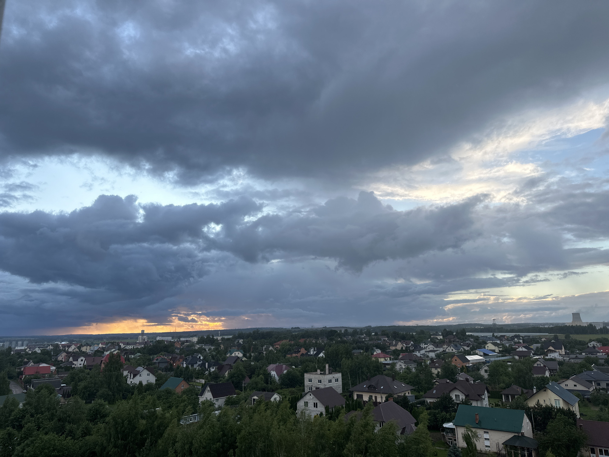 The image depicts a panoramic view of a residential area in Shchomyslitski Sel'ski Sovet, Belarus, under a dramatic, cloudy sky. The foreground is dominated by lush green trees, with their foliage creating a dense canopy. Below the trees, a multitude of houses with pitched roofs in various colors, including brown, red, white, and green, are visible. The houses are clustered together, suggesting a suburban or village setting.
In the middle ground, the houses continue to spread out, interspersed with more trees. Some larger buildings, possibly industrial or institutional, can be seen in the distance. The horizon is lined with a mixture of trees and structures.
The sky is a prominent feature, filled with heavy, dark grey clouds that suggest impending rain or a recent storm. Patches of brighter sky, tinged with yellow and orange from the setting or rising sun, break through the clouds on the left side of the image, creating a striking contrast. In the far distance on the right, industrial towers or cooling towers are visible. The overall scene conveys a sense of natural beauty combined with human habitation. No people are visible in the image.