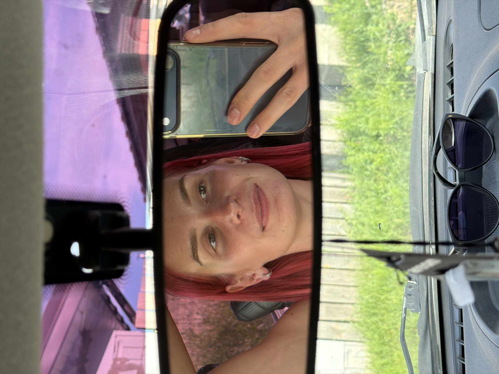 This media file captures a selfie taken in a car's rearview mirror. The primary subject is a young person with bright red hair, visible in the reflection. They are looking at the camera with a slight smile, appearing relaxed and happy. The person is wearing a silver hoop earring in their left ear.
In the reflection, a hand is holding a smartphone, also a silver color, which is clearly being used to take the picture. The background visible through the car window is a lush green outdoor scene, suggesting it is daytime with good weather.
Other objects in the car include a pair of dark sunglasses hanging on the right side of the frame, resting on a textured gray surface, likely part of the car's dashboard or interior paneling. The rearview mirror itself is framed by the car's interior, with a purple tinted windshield or tint visible on the left side of the frame, reflecting ambient light. The car appears to be parked, as indicated by the stillness of the reflection and the focus on the selfie. The overall impression is a candid moment captured during a car ride on a pleasant day.