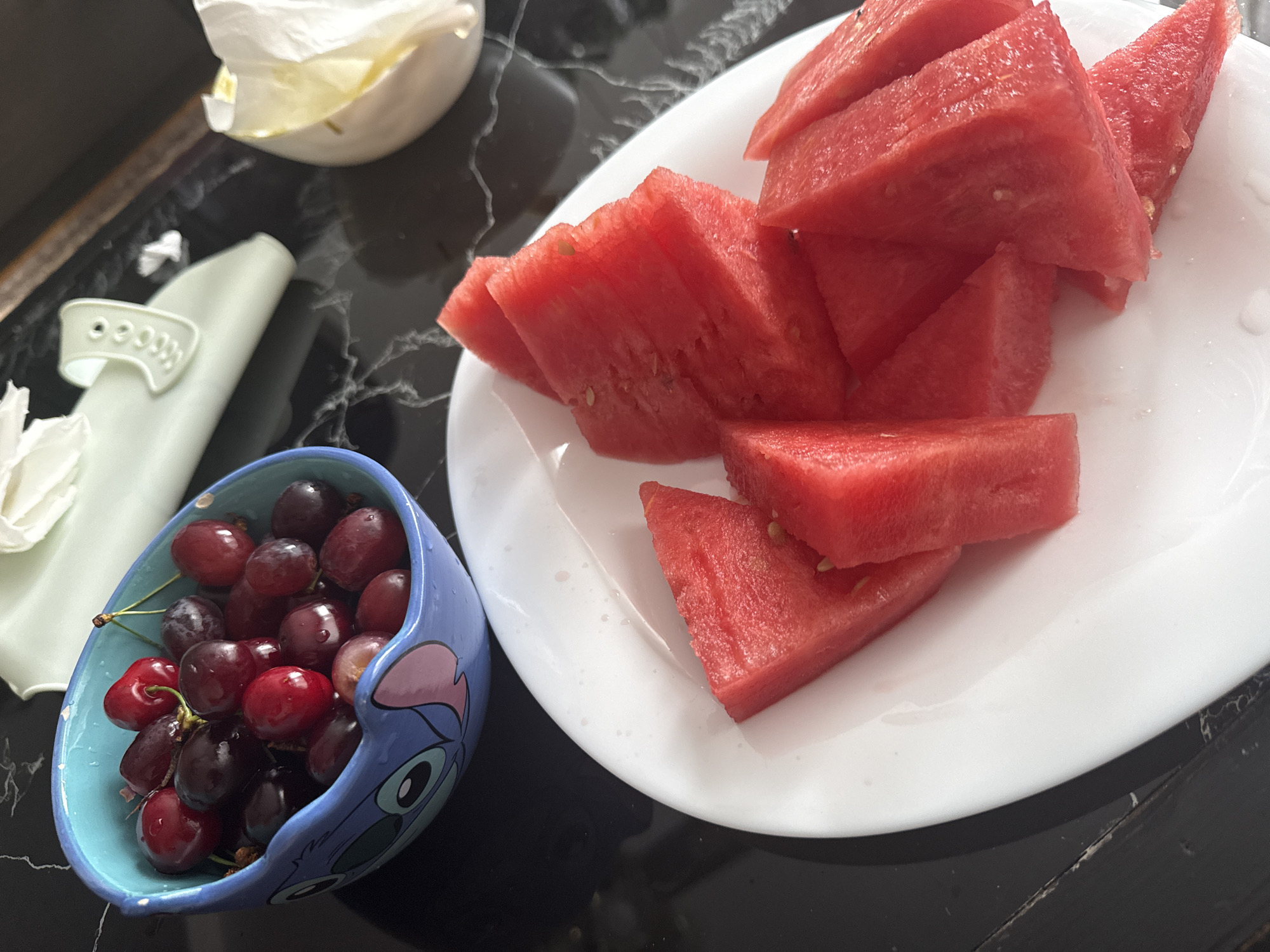 The image shows a table setting with a plate of watermelon slices and a small blue bowl of red grapes. Next to the grapes is a white cloth napkin. The table has a black, textured surface. No people are visible. The location is not identifiable as Momchilovtsi, Bulgaria based on the visual information.