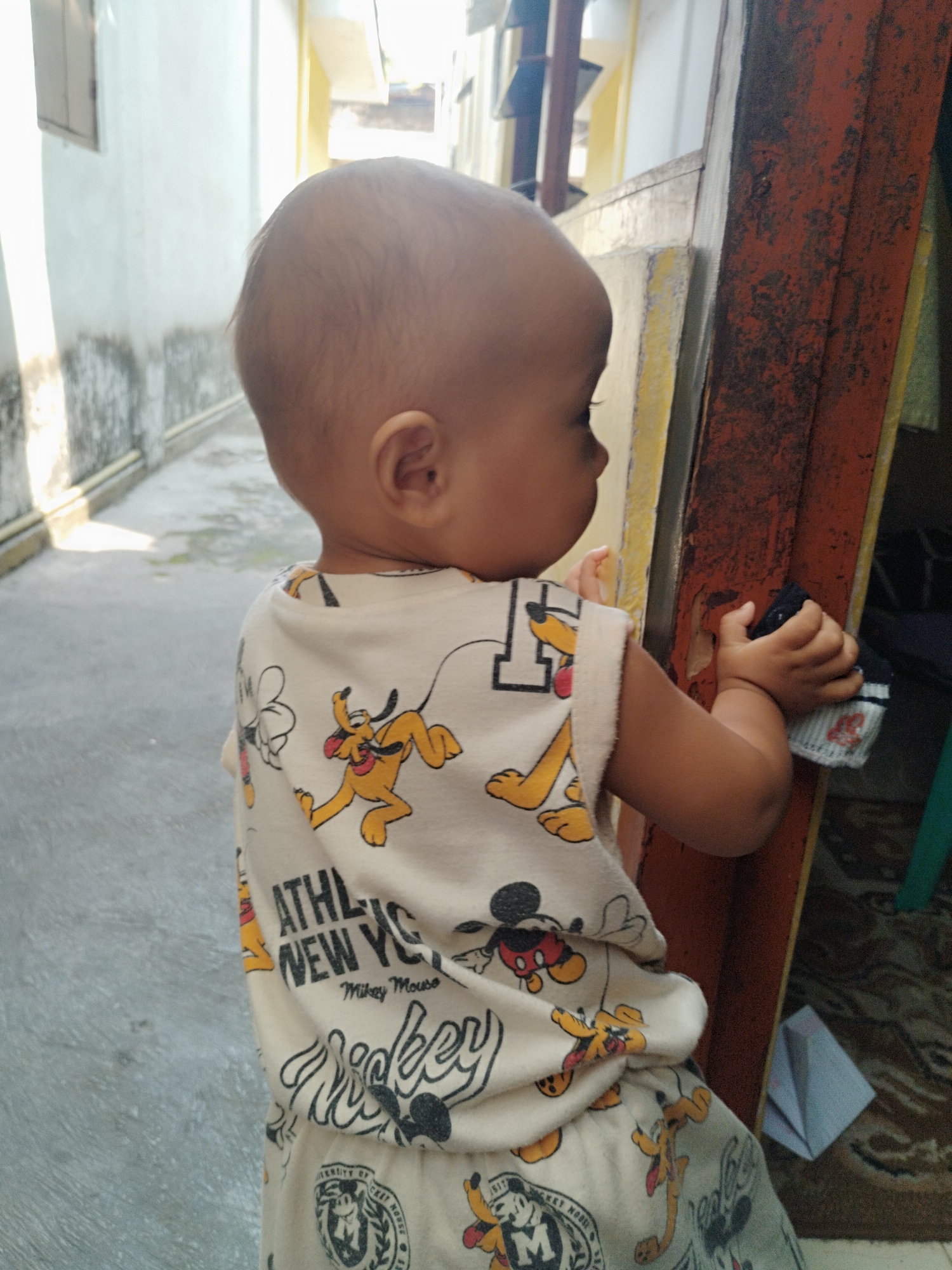 A young child, facing right and slightly away from the camera, stands in an outdoor alleyway. The child is wearing a light-colored, sleeveless shirt and pants adorned with cartoon characters, including Pluto and Mickey Mouse. The child's right hand is holding a small, dark-colored object. The alleyway features a concrete floor, a light-colored wall with some weathering on the left, and a vertical, reddish-brown wooden structure with peeling paint on the right. The background shows building structures with yellow and white elements, suggesting an urban or residential setting. The scene is illuminated by natural light, casting shadows. The location is identified as an alleyway in the city of Surabaya, Indonesia.