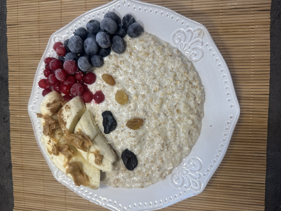 The image presents an overhead view of a white, octagonal-shaped plate filled with a breakfast meal. The plate rests on a light brown bamboo placemat, which in turn sits on a dark gray surface. The primary subject is a serving of creamy, light-colored oatmeal or porridge occupying the majority of the plate.
On the upper left side of the oatmeal, several sliced bananas are arranged, with some slices topped with light brown peanut butter and a sprinkle of reddish-brown powder, likely cinnamon. To the upper right, two distinct clusters of berries are visible: a pile of redcurrants, characterized by their bright red color and a visible layer of frost, and an adjacent pile of blueberries, also appearing frosted. Scattered across the oatmeal base are several dark raisins and lighter, golden raisins.
No people are visible. There are no discernible actions or interactions occurring. The environment appears to be an indoor setting, consistent with a kitchen or dining area. The specific location in the city of Tbilisi, Georgia, cannot be determined from the visual information provided in the image.