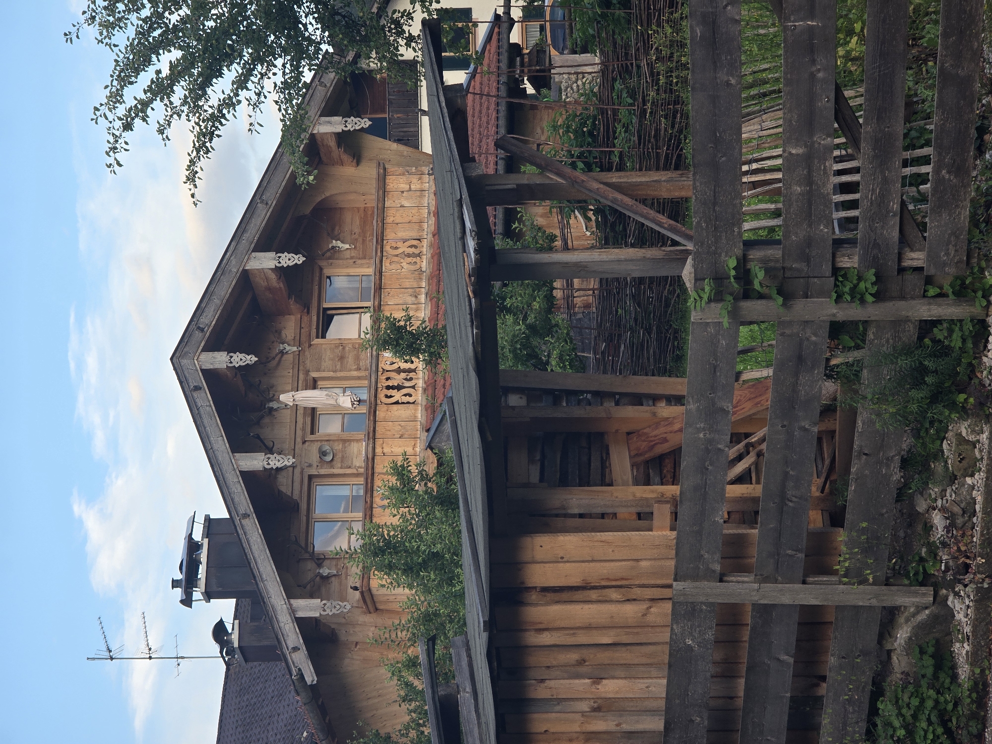 This is a vertical, eye-level, medium shot of a rustic wooden house with traditional Bavarian architectural elements, set against a blue sky with scattered clouds. The house is constructed primarily of wood, with a steeply pitched roof and decorative carvings along the eaves. Several windows are visible, some with intricate wooden shutters. Two animal skulls, likely from deer or chamois, are mounted as decorative elements below the roofline.
In the foreground, a weathered wooden fence runs across the right side of the frame, composed of vertical planks. Behind the fence, more wooden structures and fencing are visible, suggesting a yard or outdoor area. Lush green foliage, including tree branches and bushes, surrounds the house and fence, indicating a natural or semi-rural setting. The overall impression is one of traditional alpine architecture and a peaceful, possibly rural, environment. The lighting suggests it is daytime, likely late afternoon or early evening due to the warm tones on the wooden surfaces and the quality of the light. There is no visible text in the image. The scene evokes a sense of traditional charm and a connection to nature.
