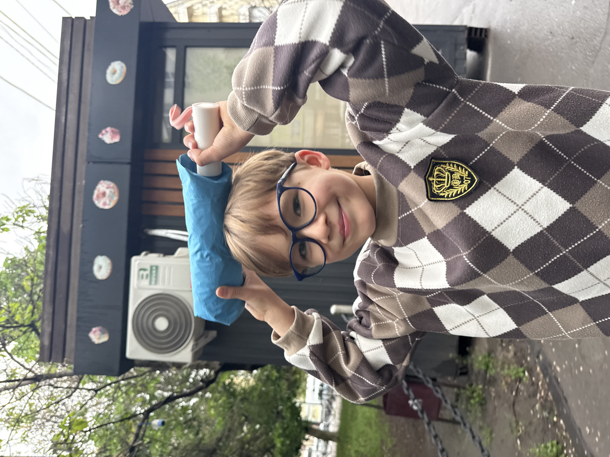 A young boy, possibly in his preteens, is standing outdoors, holding a blue, crumpled object on his head with both hands. The boy has fair skin, blond hair, and is wearing round, blue-rimmed glasses. He is smiling slightly at the camera. He is wearing a brown and white argyle sweater with a gold crest-like patch on the chest.
Behind the boy is a dark-colored building. Affixed to the building are several items: a white air conditioning unit, and what appear to be donut-shaped decorations.
The setting appears to be outdoors in a somewhat urban environment, with foliage visible at the bottom of the image. The location is Rostov-on-Don, Russia.