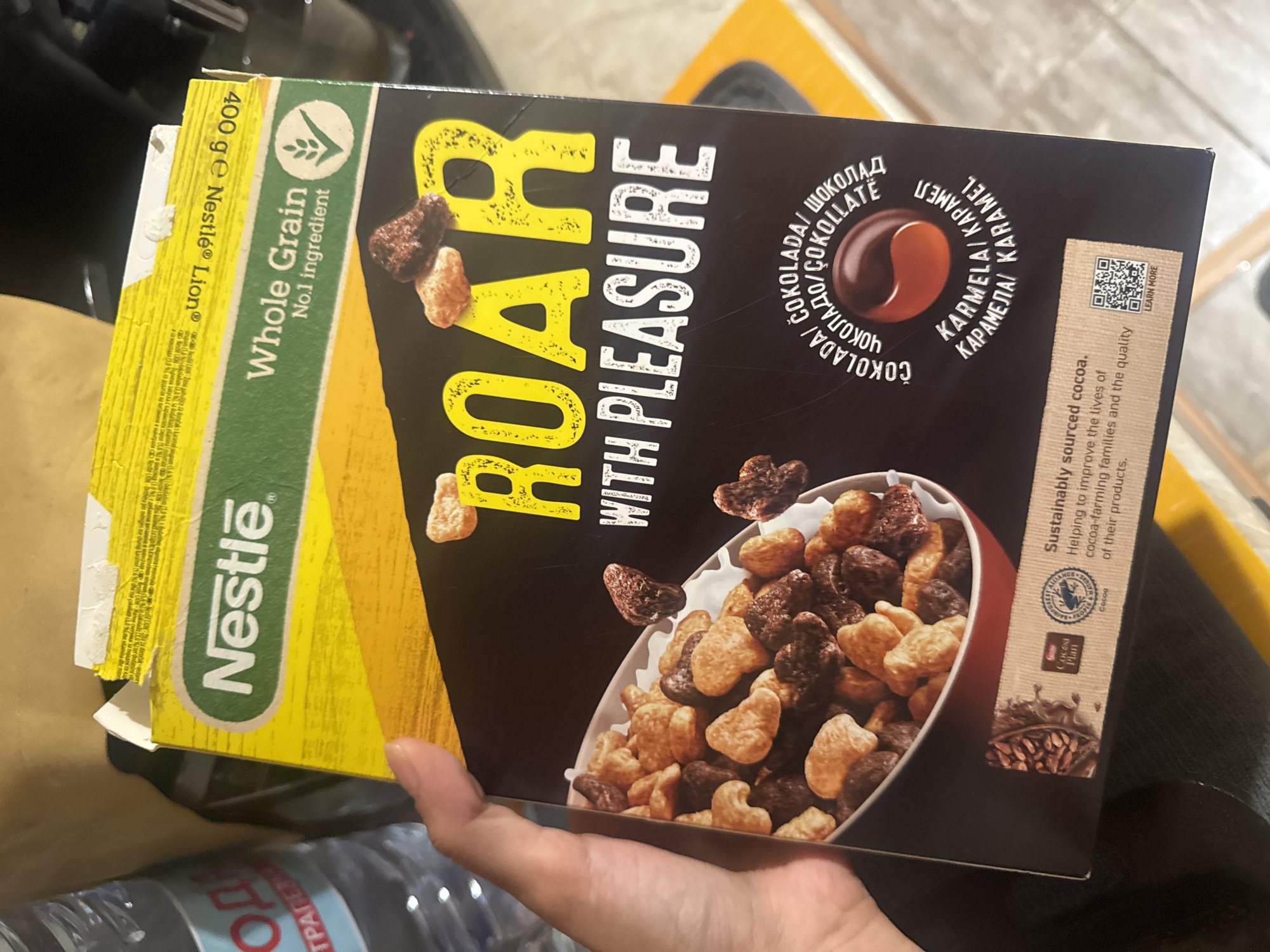 A close-up shot shows a hand holding a box of Nestle Lion cereal. The cereal box is yellow and black, with images of cereal pieces and the Nestle logo. The text on the box reads "Nestle," "Whole Grain," and "Roar with Pleasure." The background is out of focus and appears to be a domestic or retail setting.