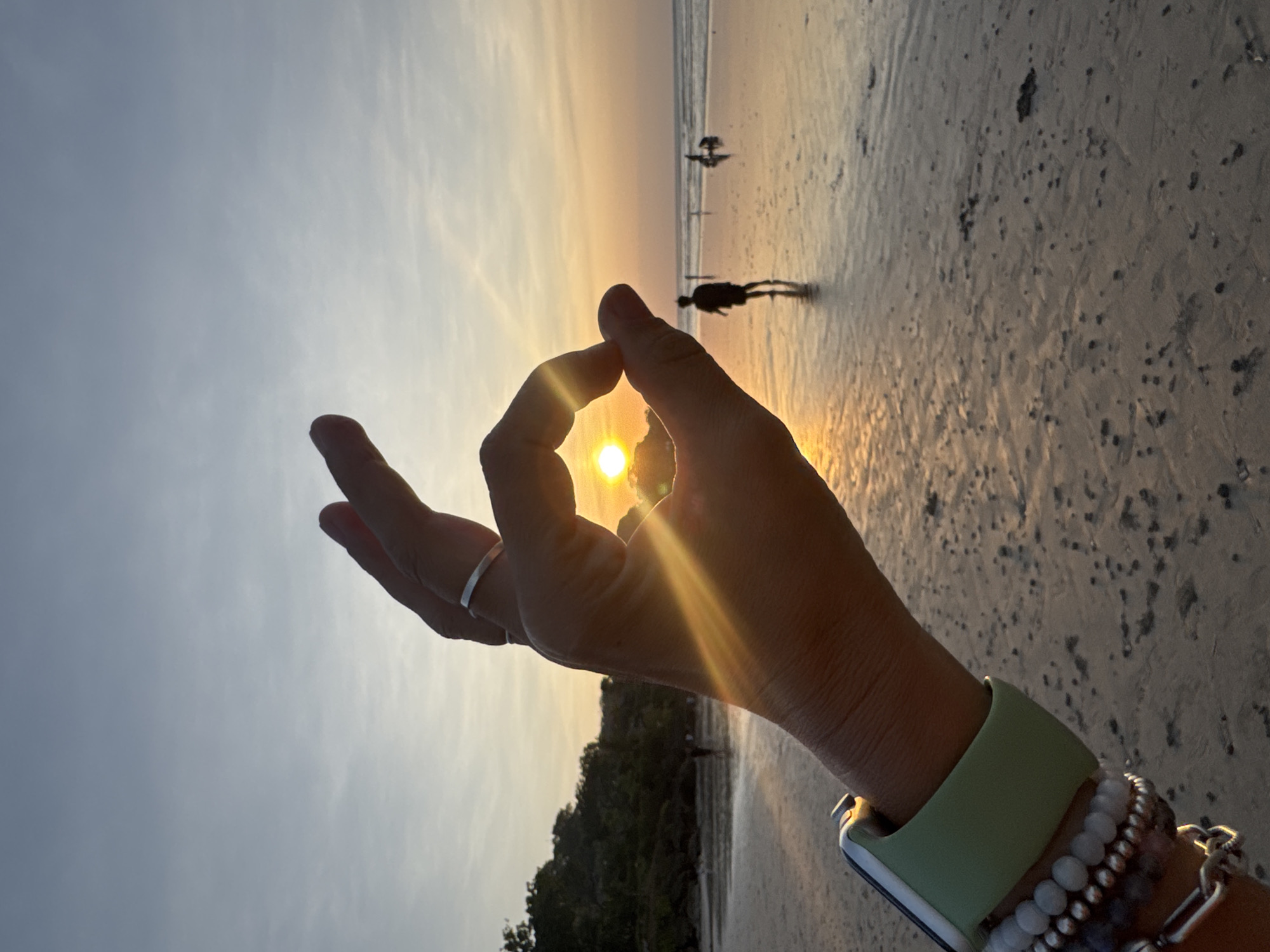 A close-up, over-the-shoulder shot captures a hand forming an "ok" symbol against a beach sunset in Kuta Selatan, Indonesia. The sun is centered within the hand's frame, its rays creating a lens flare that extends across the image. The background features a sandy beach with a few silhouetted figures walking along the shoreline and near the water's edge. Gentle waves are visible in the distance, and a tree-lined hill forms the horizon on the left. The hand is adorned with a silver ring on the index finger and a light green smartwatch band with several beaded bracelets on the wrist. The sky is a gradient of pale blue to soft orange, indicative of either sunrise or sunset. The sand on the beach is wet and reflects the warm light.