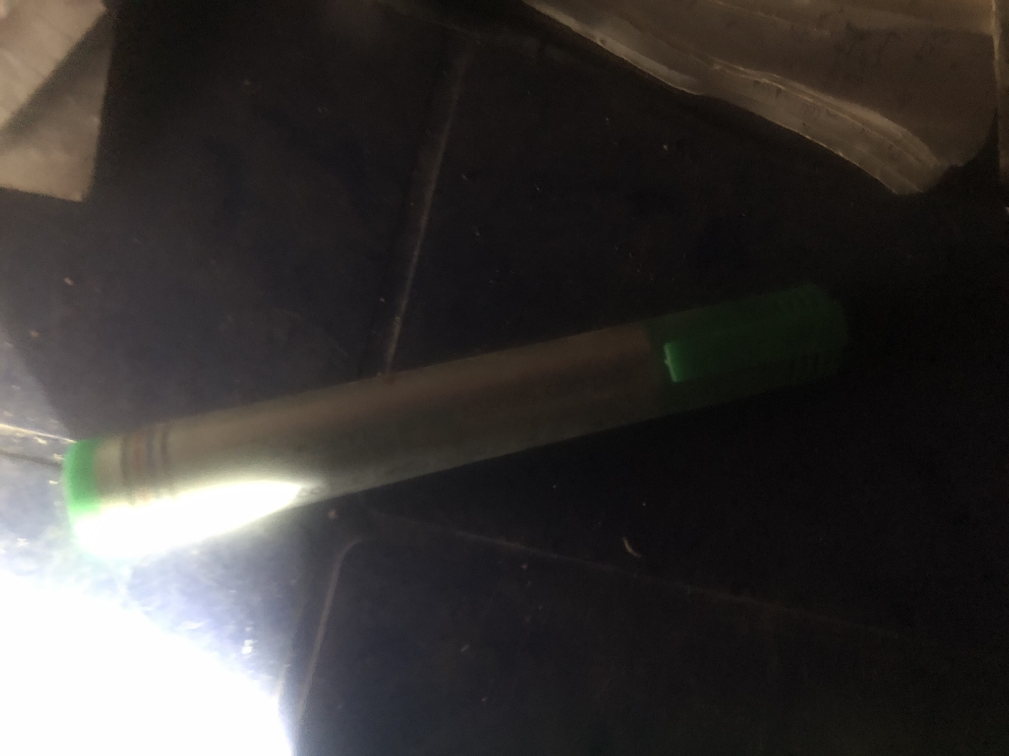 A long, cylindrical object, appearing to be a marker or pen, rests diagonally on a dark, textured surface. The object has a metallic or silver-colored body with a bright green cap or tip at the bottom. The top of the object is obscured by a bright light source, creating a significant glare and obscuring details in that area. The surface beneath the object is dark and appears to be made of wood or a similar material, with some visible dust or debris. To the right of the object, a lighter-colored, textured material, possibly paper or fabric, is partially visible. The overall lighting of the scene is uneven, with the bright light source dominating the top left portion of the image and casting shadows. There are no people or animals visible in the image, and no specific location in Wukari, Nigeria, can be identified from the provided visual information.
