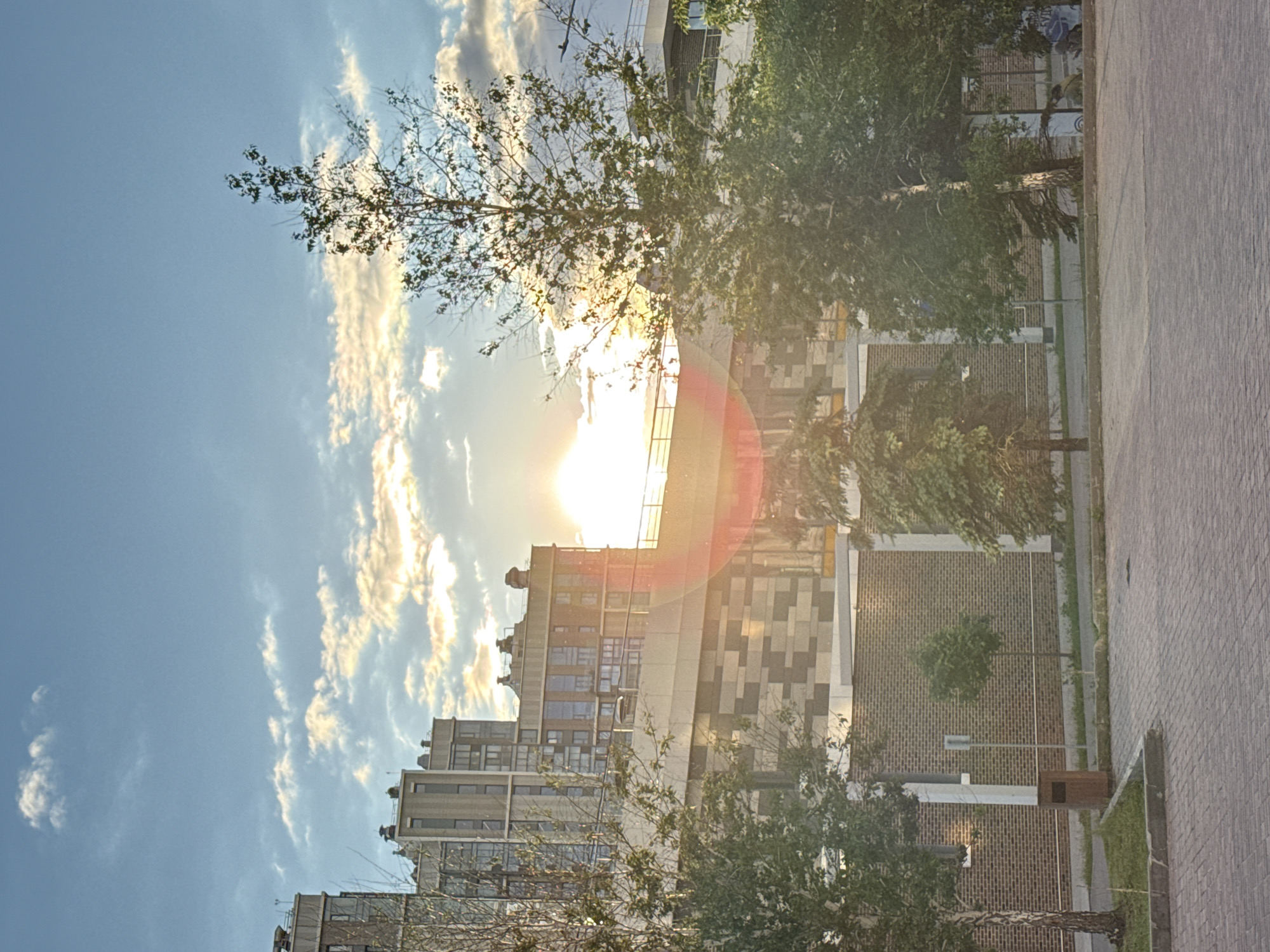 The image depicts a cityscape at sunset, likely in Moscow, Russia, given the architectural style of the buildings. The sun is positioned centrally, creating a lens flare effect. The foreground features trees with leaves silhouetted against the bright light. Behind the trees, a multi-story building complex is visible, characterized by a mix of brick and concrete facades with a grid-like pattern. The sky is blue with scattered white clouds, some illuminated by the setting sun. A paved walkway is visible on the right side of the image.