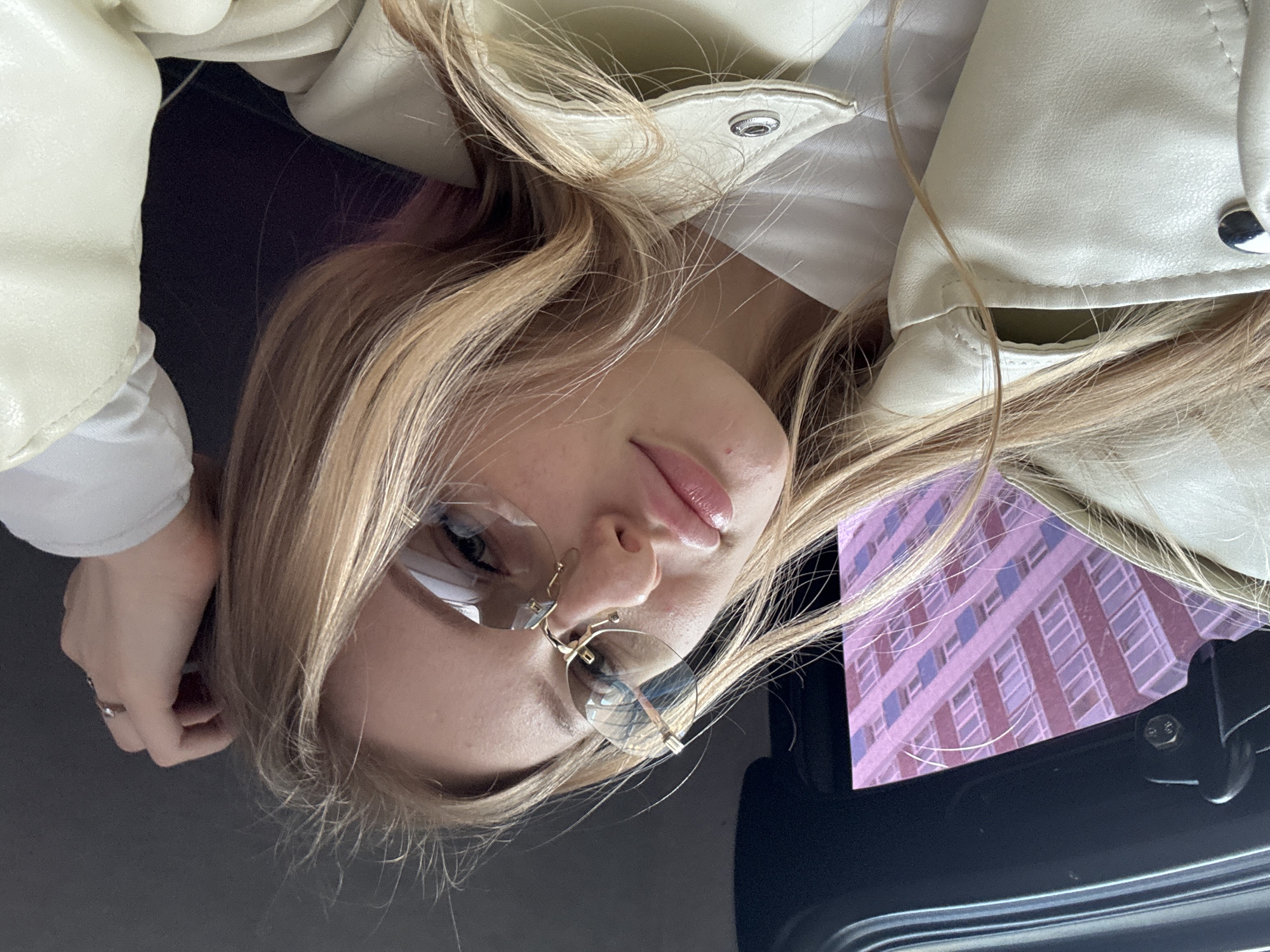 The image shows a person with blond hair wearing a light-colored jacket and glasses, likely in a car. The reflection in the car window shows a building with a grid-like pattern, suggesting the city of Perm, Russia. The person is looking down at the camera. Their left hand is visible with a ring.