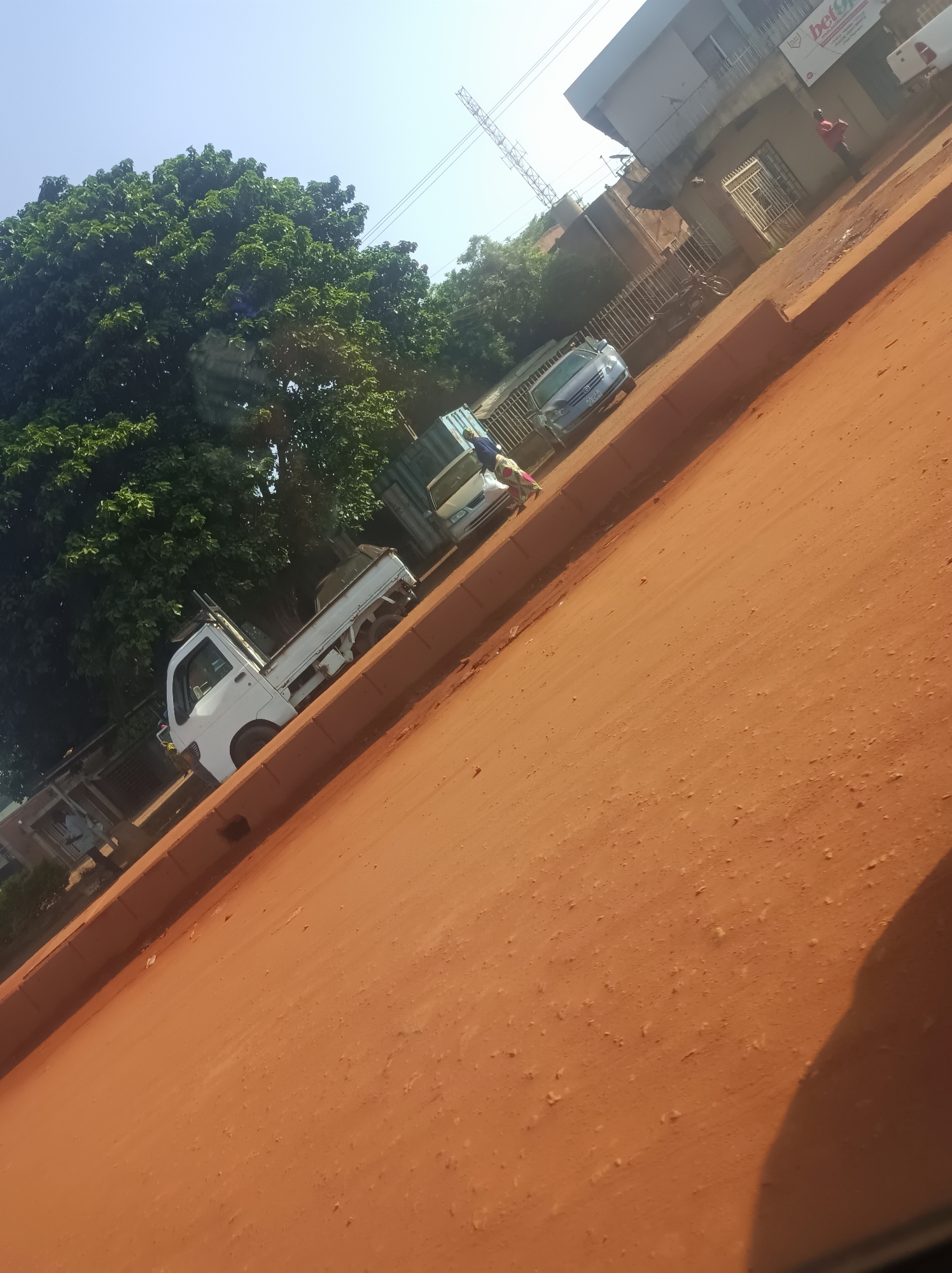 The image captures an outdoor scene, likely a street or unpaved road in Barnawa, Nigeria, during the daytime under a clear, bright sky. The dominant feature is a wide expanse of reddish-brown dirt, characteristic of laterite soil, which forms the road and shoulder. A concrete curb separates this dirt area from the side of the road where buildings and vehicles are located.
On the left side of the image, a large, dense green tree with abundant foliage occupies the upper portion, extending down to frame parked vehicles. A white compact pickup truck with an open bed is prominently visible, positioned close to the curb. Further along, a light-colored sedan or small SUV is also parked. A person wearing patterned clothing is partially visible near the white truck.
In the mid-ground, a two-story building with a light-colored facade is visible. A sign on this building clearly displays "bet9ja" in white text on a red background, indicating a betting shop. Below it, smaller text likely details services. A person wearing a red shirt is standing near this building. To the left of the "bet9ja" building, another structure and a metal fence are visible. Further down the road, a blue vehicle, possibly a container truck, is parked, with another person in blue visible near it.
The overall impression is a bustling local street scene, with parked vehicles, people present, and commercial activity suggested by the "bet9ja" sign. The ground appears dry and dusty. A dark shadow partially obscures the bottom right corner, suggesting the photo was taken from inside a vehicle.