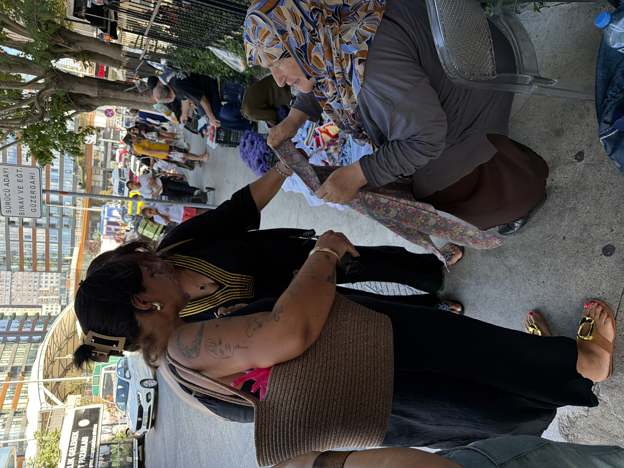 In the city of Kadıköy, Turkey, on a sidewalk bordered by trees and a black metal fence, two women are engaged in a transaction. One woman, wearing a black dress, a prominent gold necklace with a black pendant, and gold hoop earrings, holds a woven brown bag. She is reaching out to another woman, who is seated on a transparent plastic chair. This seated woman wears a gray top and a dark brown skirt, with her head covered by a patterned hijab featuring floral designs in blue, brown, and yellow. She is holding a colorful floral fabric, appearing to be a scarf or textile, and is showing it to the standing woman.
Behind the seated woman, stacks of purple and white fabric are visible, along with other items on the ground. To the left of the main interaction, a large white bus with Turkish text is parked on the street. Further down the street, several cars and pedestrians are present. The background shows modern buildings. The overall scene depicts a street market or a vendor interacting with a potential customer on a sunny day.