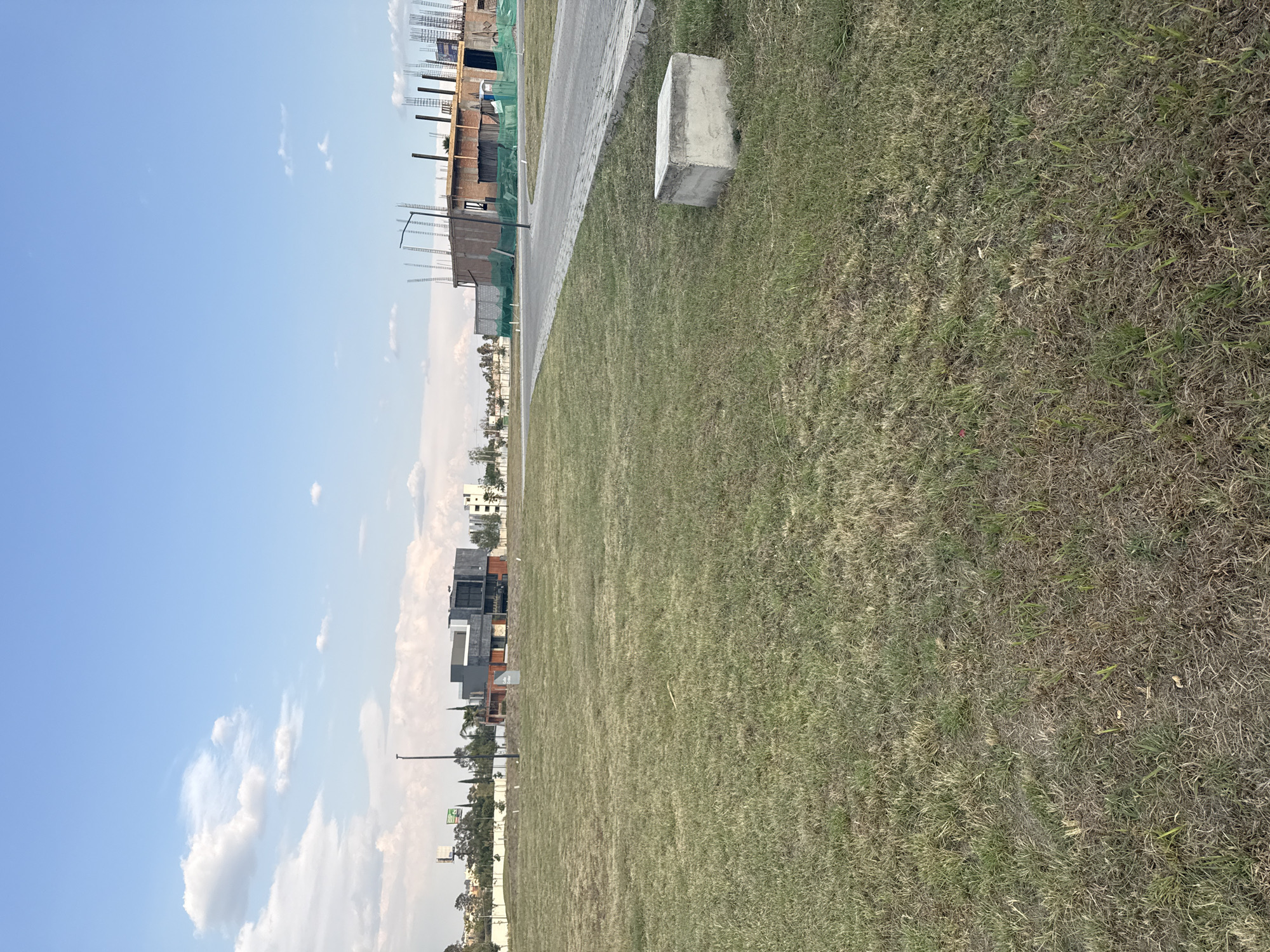 The image shows a grassy area with a concrete curb or sidewalk and a concrete block. In the background, there are buildings under construction, and a few completed modern houses. The sky is light blue with some clouds. The image location is San Bernardino Tlaxcalancingo, Mexico. There are no people visible in the image.