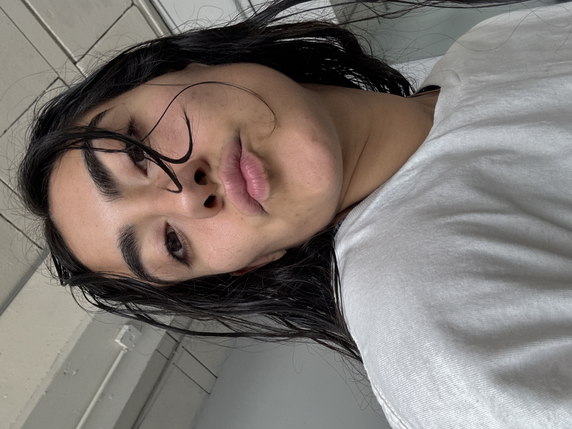 The scene depicts a close-up, low-angle portrait of a single female subject in an indoor setting, stated to be in the city of Puebla, Mexico. The subject has dark, wet hair, with several strands adhering to her forehead and right cheek. Her skin tone is fair to olive, with visible pores. She possesses prominent dark eyebrows and dark brown eyes, which are directed generally towards the viewer. Her lips are pursed in a distinct "kissing" gesture. She is attired in a plain white t-shirt, exhibiting a subtle ribbed texture along the neckline and shoulder area. The background primarily consists of a plain white ceiling, featuring visible white electrical conduits or pipes and a white electrical box. The illumination is consistent with ambient indoor lighting. No other individuals or interactions are present.