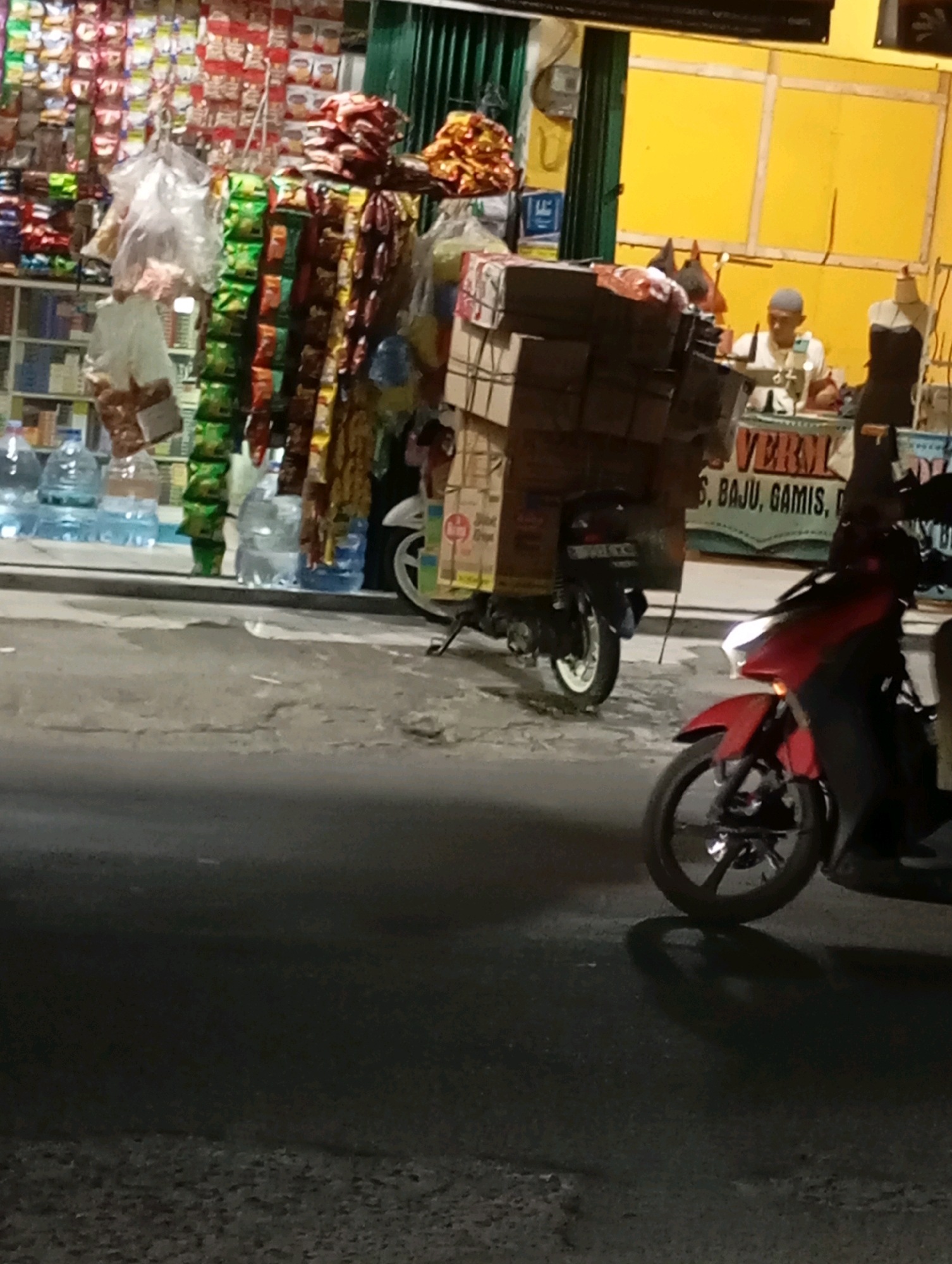 The image depicts a nighttime street scene in Babelan, Indonesia, in front of a storefront displaying various goods. A person is seated at a counter, and several motorcycles are parked or in motion.
In the foreground, a red motorcycle is parked, its headlight illuminated. Its front wheel and part of its frame are visible. The road surface appears to be asphalt with some debris.
In the middle ground, a black motorcycle is loaded with a large stack of boxes, suggesting it is used for transport. The boxes are strapped to the rear of the motorcycle. To the left of this motorcycle, several large bottles of water are arranged on the ground.
Behind the motorcycles, the storefront is visible. It is cluttered with various items, including bags of snacks or goods hanging from the ceiling, and shelves filled with products. The storefront has a yellow wall in the background. A person wearing a light-colored shirt and a cap is seated at a counter within the store, possibly attending to customers or engaged in work. A mannequin dressed in dark clothing stands to the right of the counter. A sign with text, possibly advertising goods, is partially visible behind the counter. The overall impression is a busy, local shop operating at night.