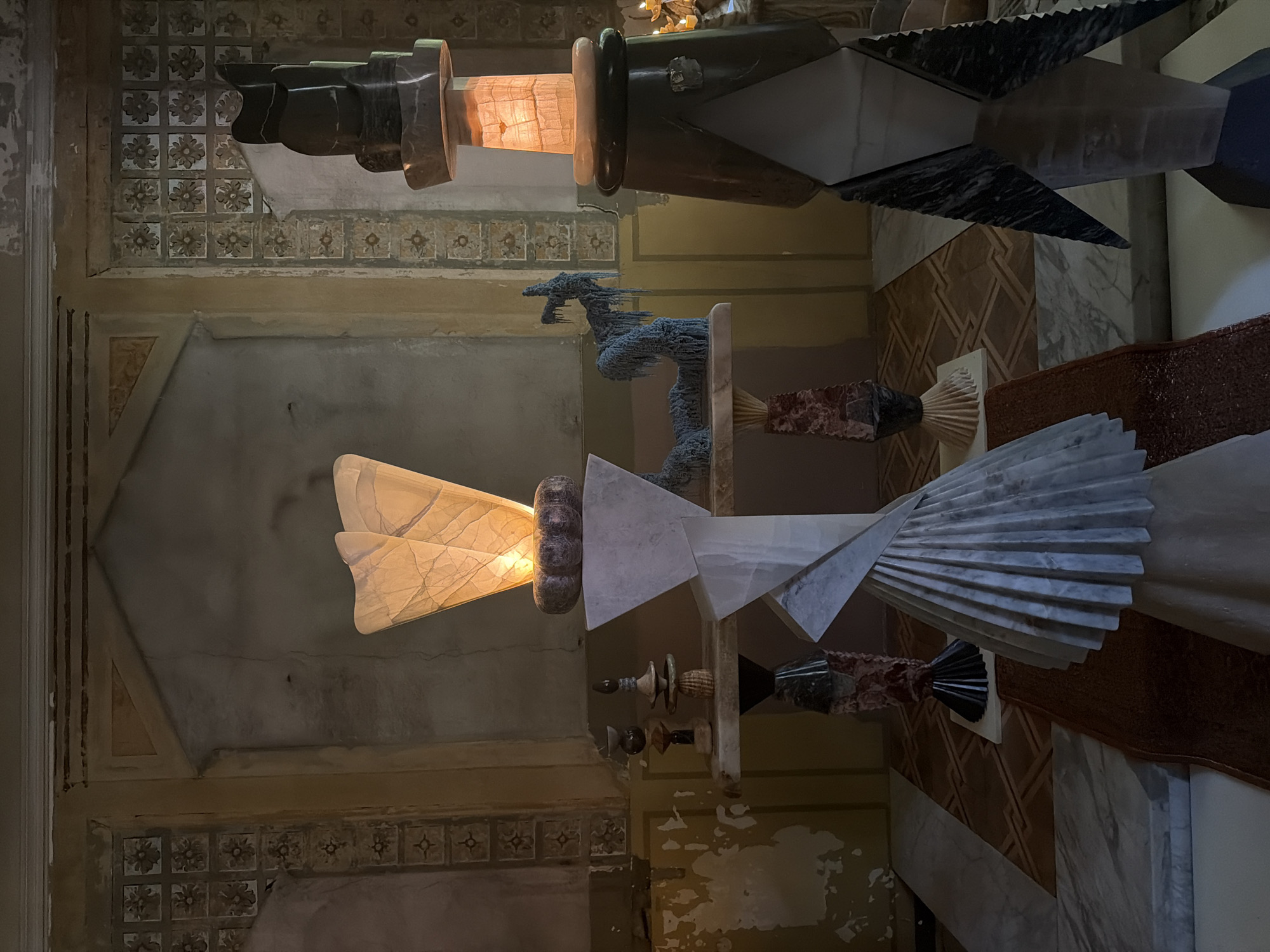 The image shows the interior of an ornate room in Milan, Italy, likely a gallery or museum exhibiting contemporary art. Two large-scale sculptures dominate the frame. The upper sculpture is composed of dark stone and a section of translucent material that is glowing with light. The lower sculpture consists of white and gray stone, with a similar translucent section. A metal dragon sculpture sits between the two. The room has patterned walls and marble flooring. The lighting is a combination of natural and artificial, with areas of shadow and highlight. There are no people visible in the frame.