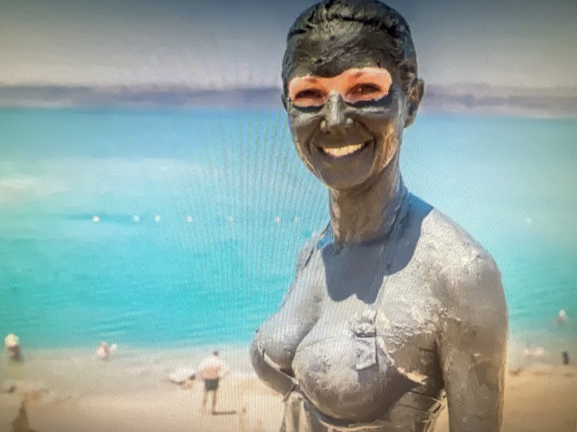 A person with their face and body covered in dark mud is standing on a sandy beach. The mud forms a mask around their eyes, leaving the eyelids and a portion of the surrounding skin visible and exposed. The person is smiling broadly. They are wearing a light-colored garment that is also partially covered in mud. The background shows a bright turquoise body of water with a hazy horizon. There are a few other people visible in the water and on the shore, some of whom appear to be swimming or wading. The overall setting suggests a recreational area at a body of water. The location is not the city of Cheb, Czechia.