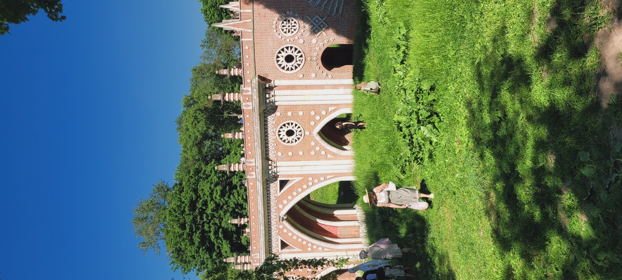 The image displays a sunlit outdoor scene at the Tsaritsyno Museum-Reserve in Moscow, Russia. The central feature is a large, ornate brick bridge structure, consistent with the Figured Bridge or Large Bridge, characterized by its red brick, white architectural accents, and Gothic Revival style. This structure presents multiple pointed arches and circular window-like openings with spoke patterns, along with decorative columns, moldings, and numerous finials crowning its upper railing. Behind and above the brickwork, a dense expanse of green deciduous trees is visible, set against a clear, deep blue sky.
The foreground and mid-ground consist of a lush green grassy area, showing variations in height and containing broader-leafed plants. Patches of bare earth are visible within the grass, particularly in the lower foreground. Several individuals are present on the ground level. In the central foreground, a woman wearing a light-colored, possibly striped dress and a wide-brimmed straw hat walks towards the right, holding a light-colored bag in each hand. To her left, near the bridge's archways, a person with grey hair, dressed in a light-colored patterned garment, is observed from behind, holding a pink bag; a small child in a light dress stands nearby. Further left, a partial view of another individual shows them carrying a long, light-colored object. Under an archway on the right side of the bridge, a woman in a dark dress stands, facing right. To her right, a person in a light-colored patterned dress kneels or squats in the grass. Additionally, several small figures, identified as people, are discernible walking or standing on the elevated top surface of the bridge. The scene is illuminated by bright, direct sunlight, creating distinct shadows on the grassy ground in the lower portion of the image.
