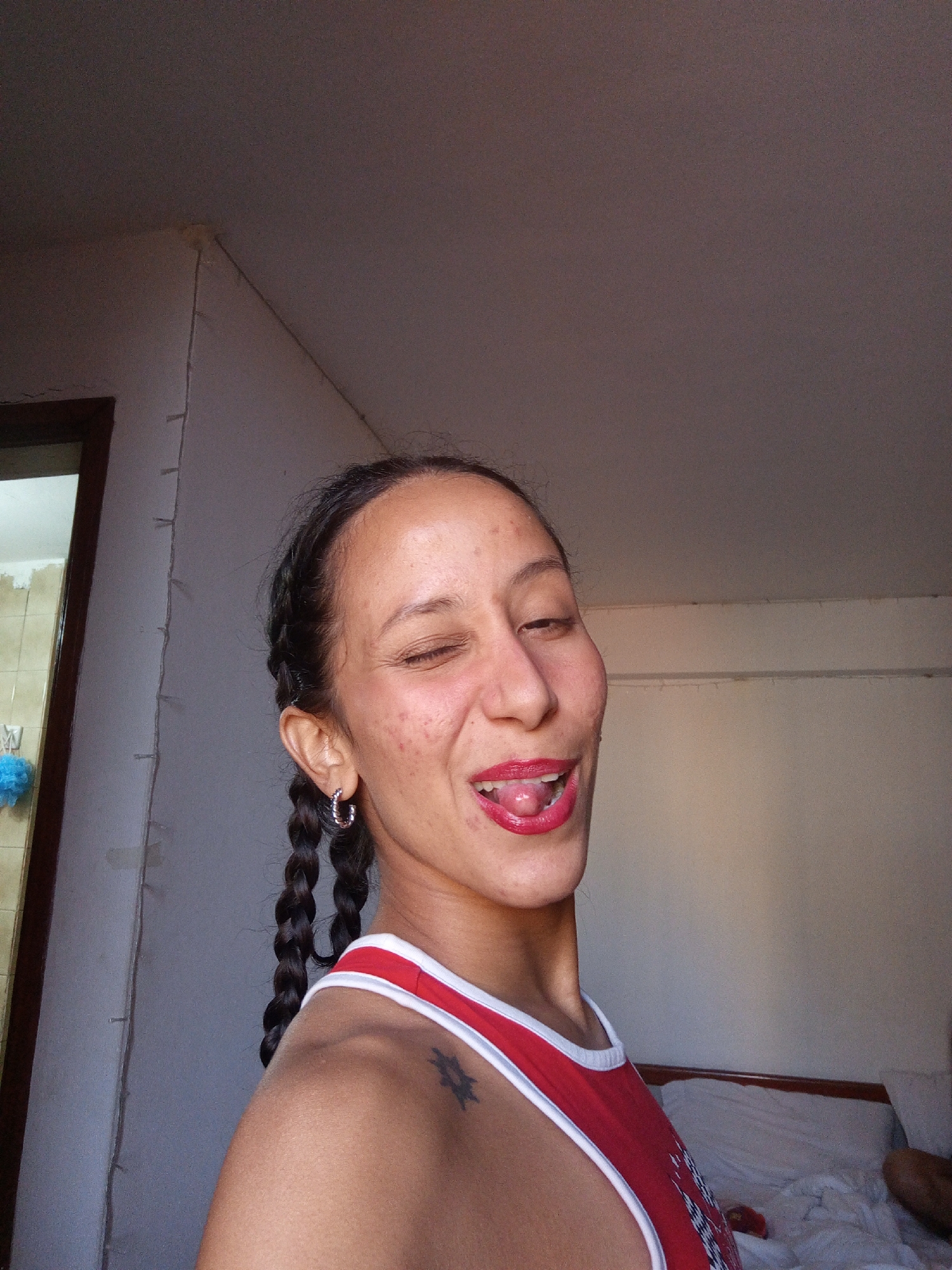 A woman is posed in a selfie, taken indoors with natural light. She has dark braided hair, is wearing a red and white tank top, and has hoop earrings. Her mouth is open, with her tongue sticking out, and one eye is winking. A faint tattoo is visible on her left shoulder. The background shows a wall and a bed. The setting appears to be a bedroom in Porlamar, Venezuela.