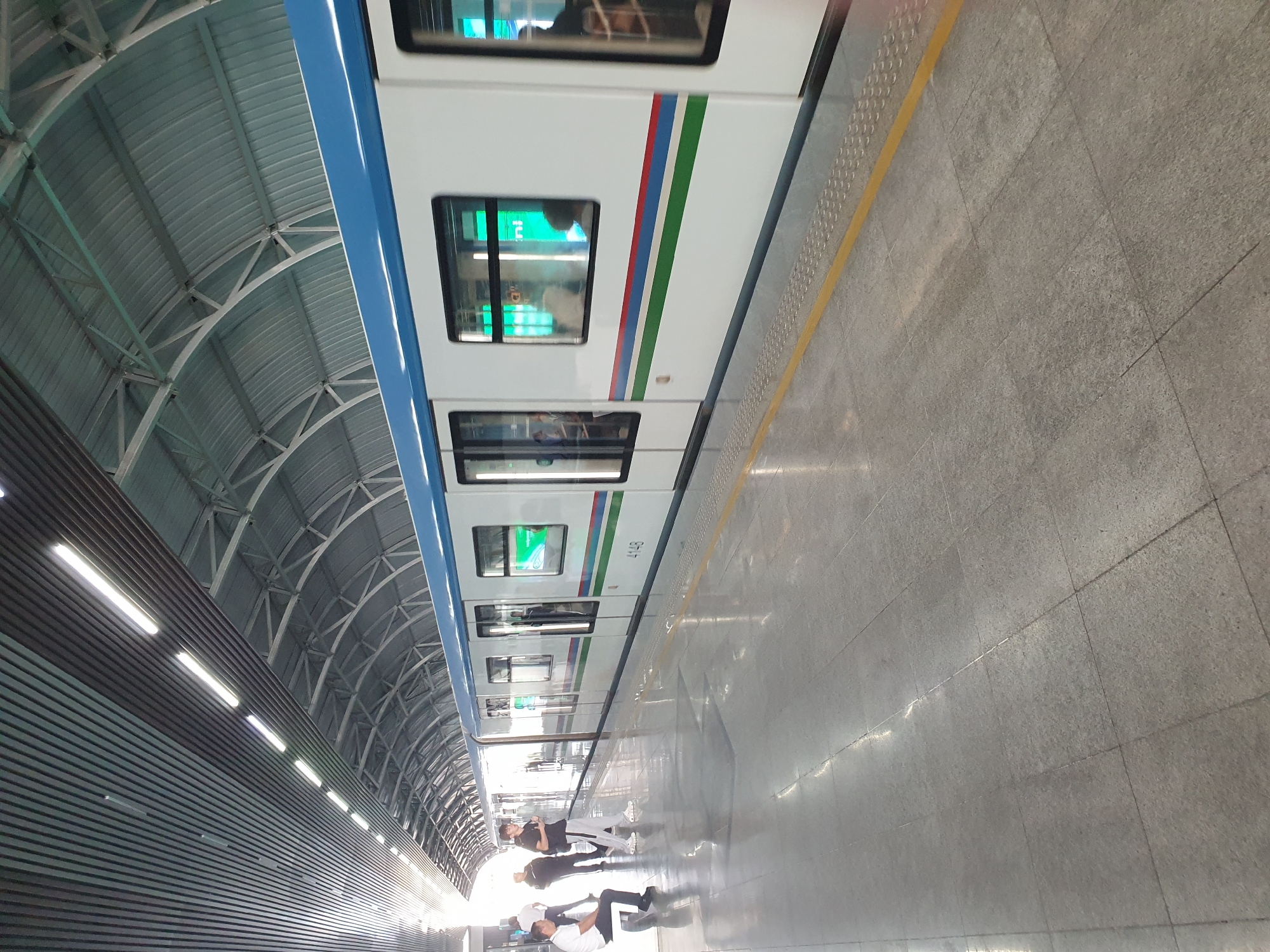 The image depicts a metro train at a station in Tashkent, Uzbekistan. The train is white with a blue stripe along the top and green, red, and blue stripes along its side. The number "4148" is visible on the side of the train. The station platform is made of grey tiles with yellow tactile paving along the edge. People are visible on the platform, some standing and some sitting. The station has a modern, covered roof structure with metal beams and corrugated metal sheeting. The lighting in the station appears to be from overhead fluorescent tubes and natural light filtering in from the open ends of the station. The scene suggests a typical operational day at a metro station.