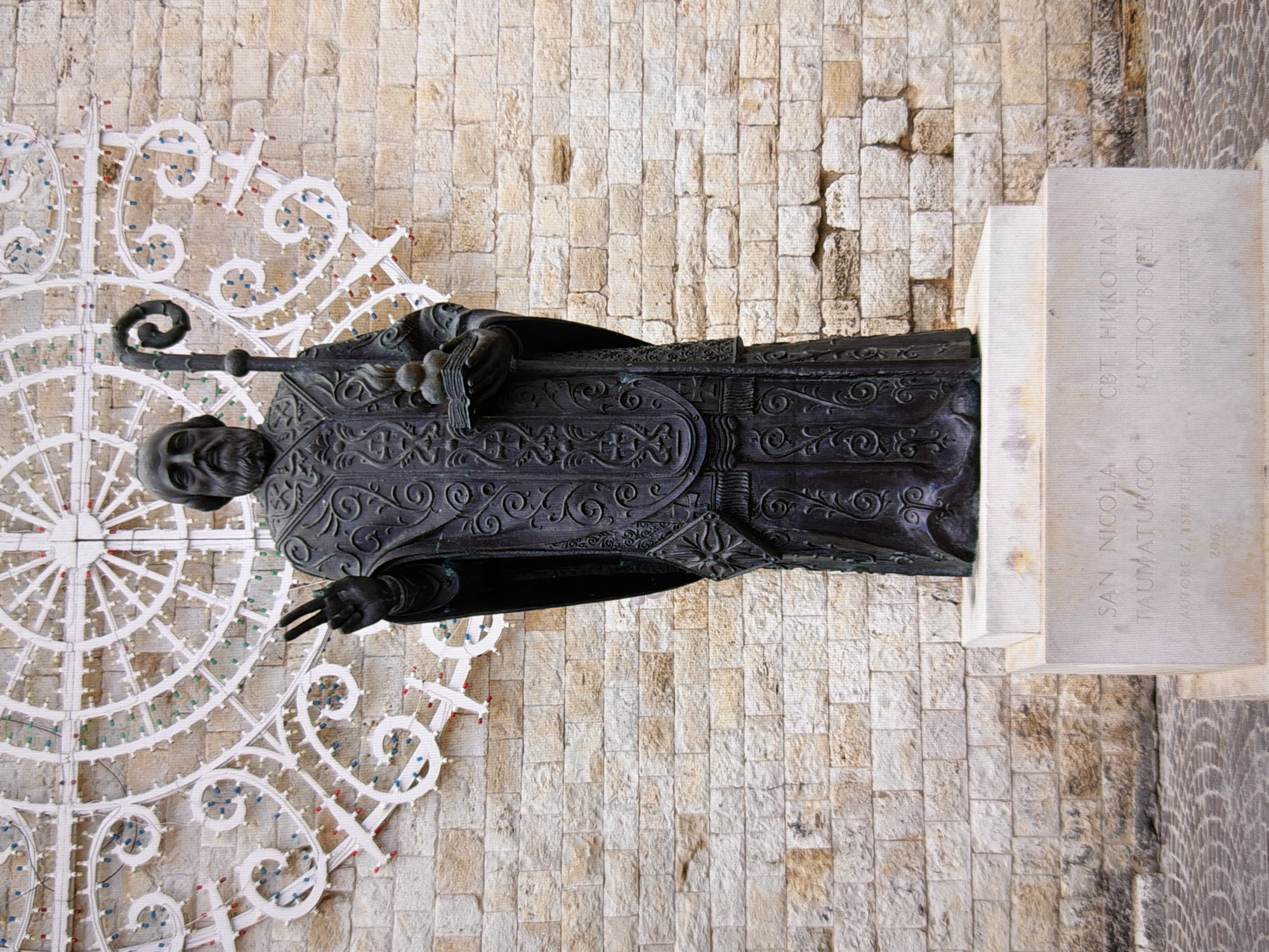 The image shows a bronze statue of Saint Nicholas, identified by the inscription "SAN NICOLA TAUMATURGO" and "СВТ. НИКОЛАИ ЧУДОТВОРЕЦ". The statue depicts Saint Nicholas in religious vestments, holding a staff in his left hand and a book with three spheres on top in his right hand. He is making a blessing gesture with his right hand. The statue stands on a stone pedestal, which bears further inscriptions including the artist's name "AUTORE Z. TSERETELI" and the year "2003". Behind the statue, a wall constructed of large, rough-hewn stone blocks serves as the background. Above the wall, an intricate white metal decorative element, resembling a large lace-like circular motif adorned with numerous small colored lights, is visible. The ground in front of the pedestal is paved with cobblestones. The lighting suggests it is daytime, with the statue and its surroundings illuminated by natural light. The overall scene appears to be an outdoor public space.